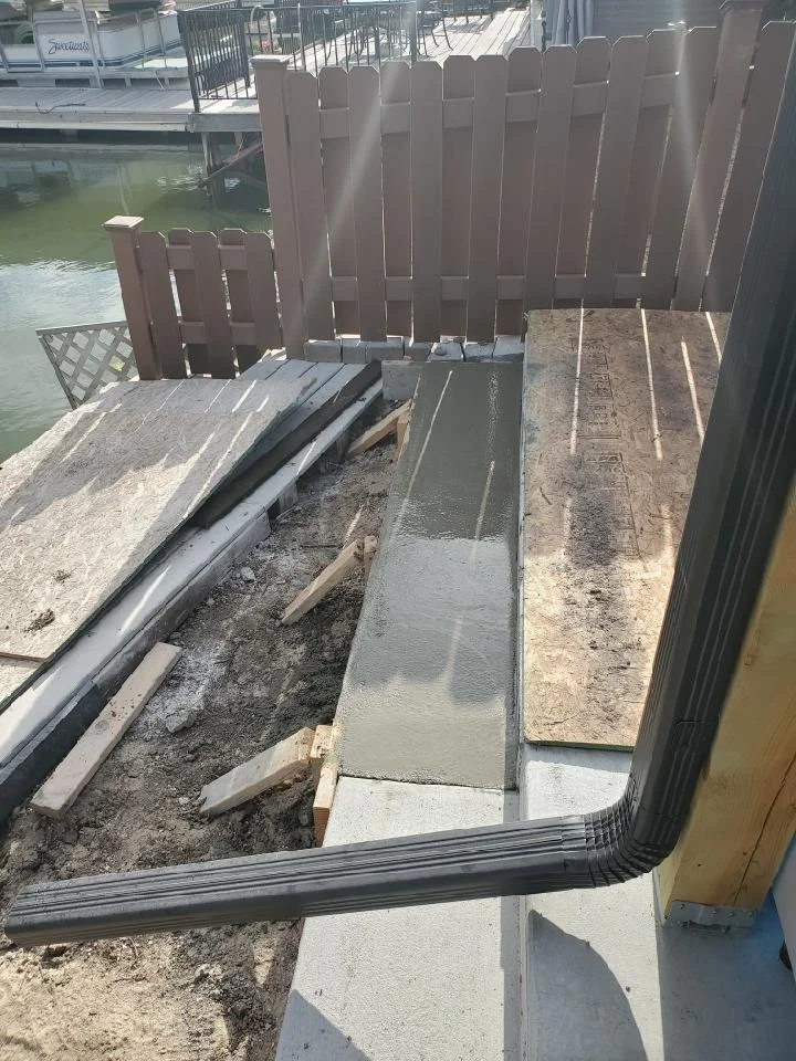 Partially finished outdoor construction area near a waterfront with a concrete pathway and a black drainage pipe.