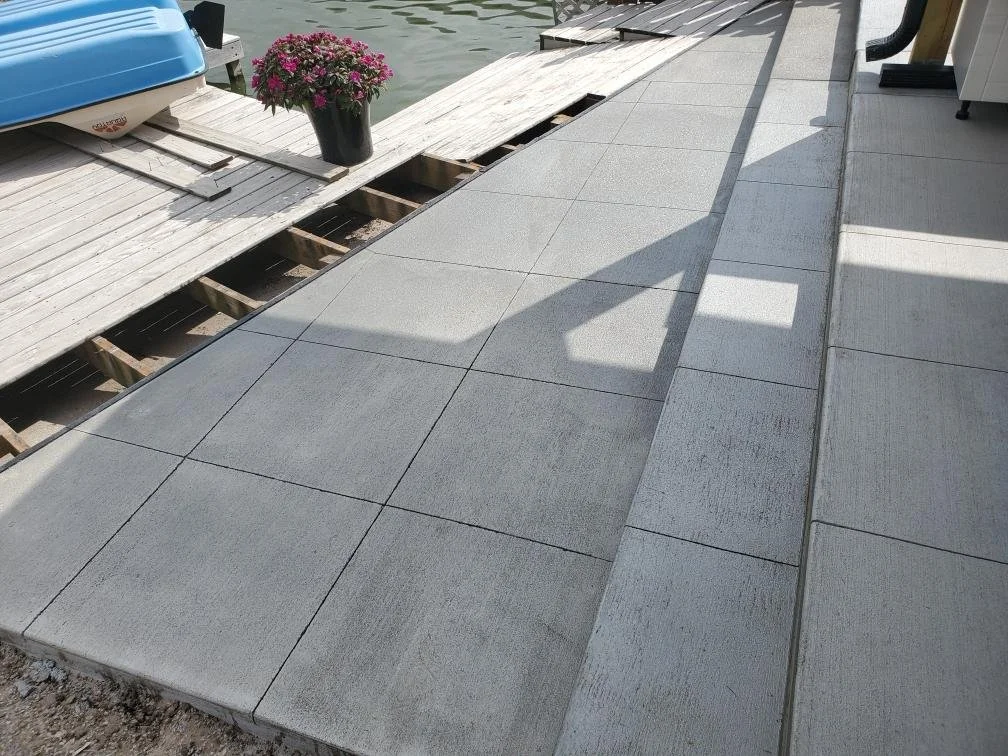 Newly installed concrete tiles on a patio or walkway beside a body of water with a shadow cast on the tiles. There is a potted plant with pink flowers and a dock with a boat in the background.