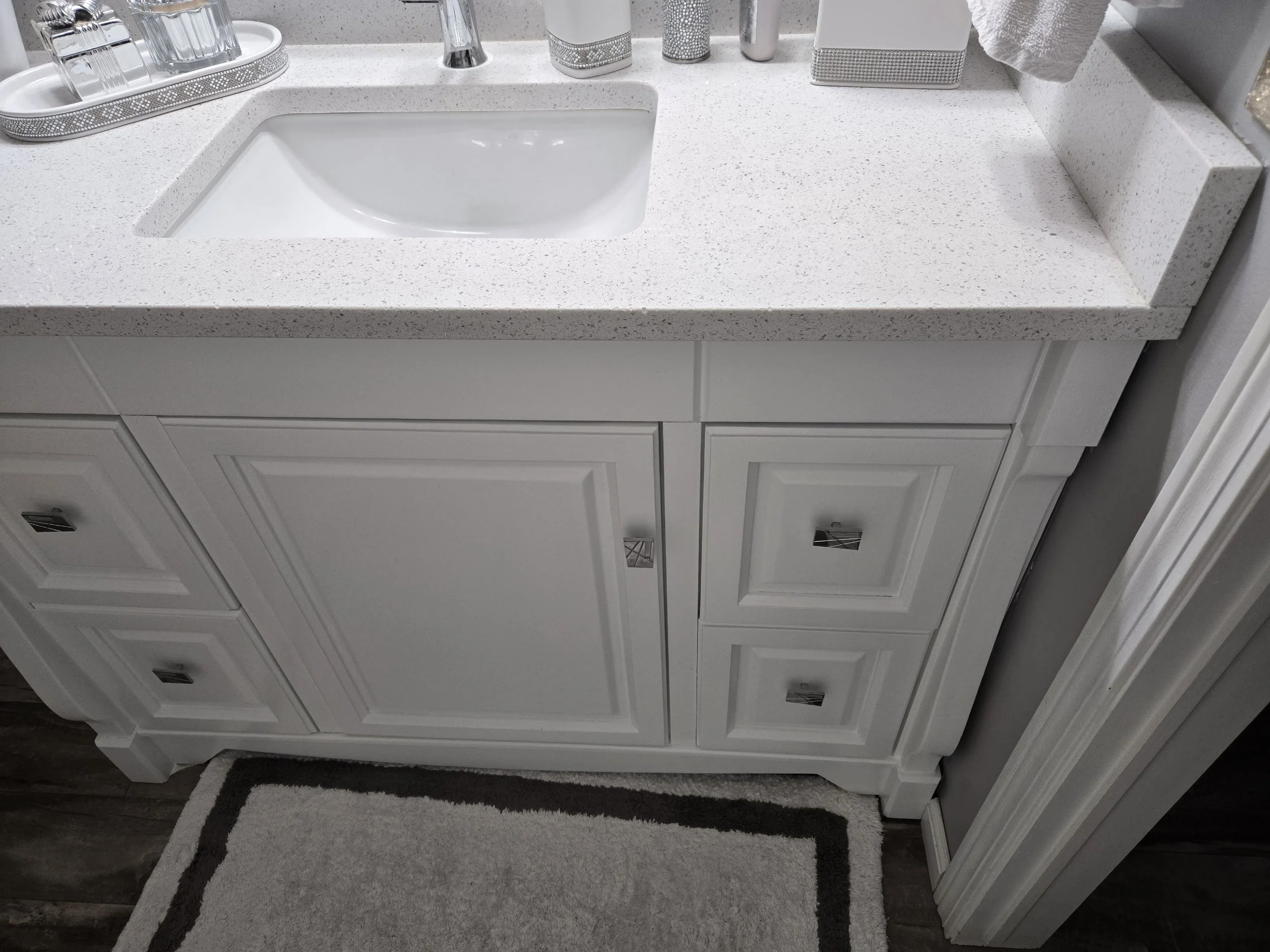 White bathroom vanity with a speckled white countertop, a built-in sink, silver faucet, and storage drawers with square silver handles. Various bathroom accessories on the countertop, including a tray with glassware, soap dispenser, and decorative it