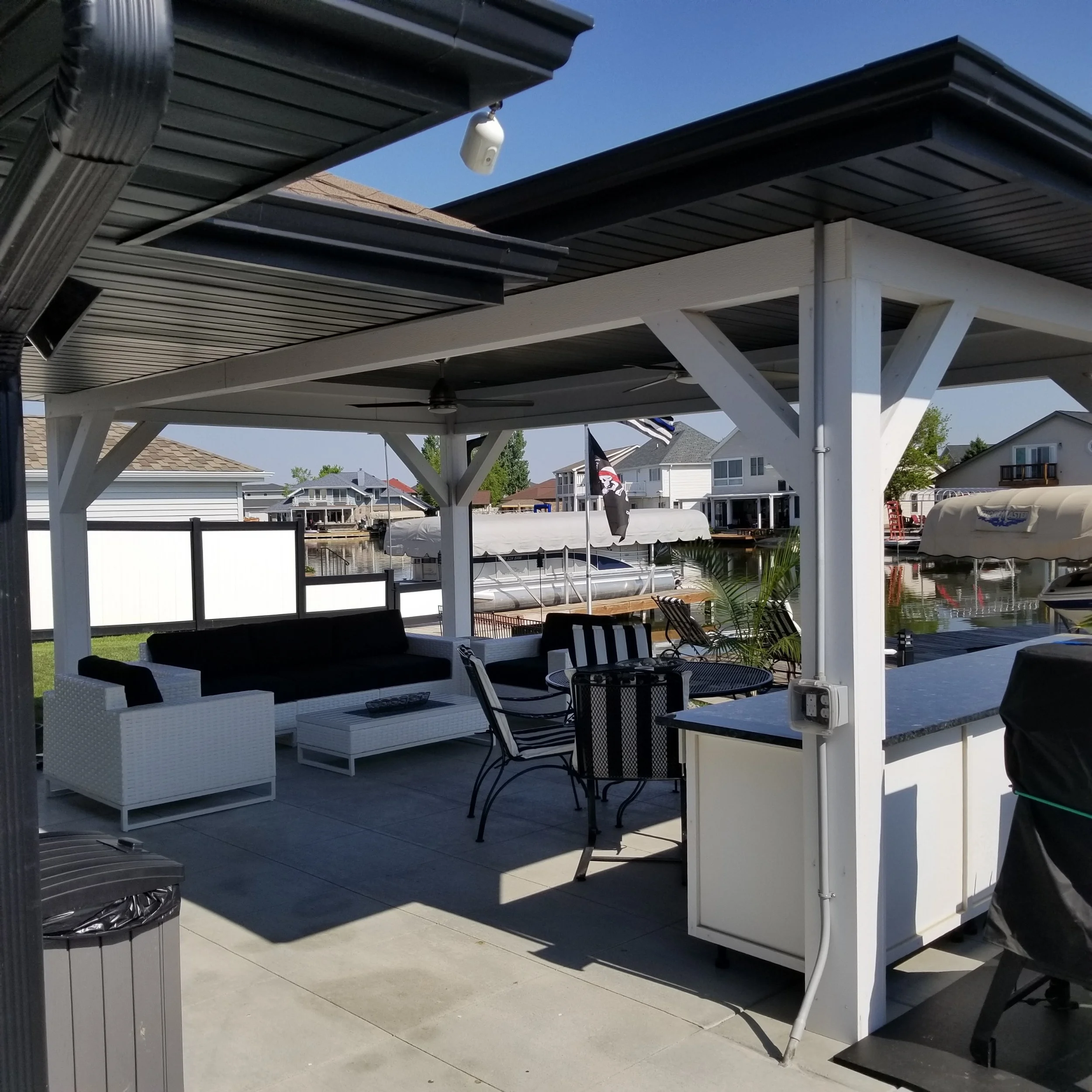 Outdoor patio with black and white furniture, including a black sofa, white armchair, striped chairs, and a bar counter, overlooking a canal with boats and houses.