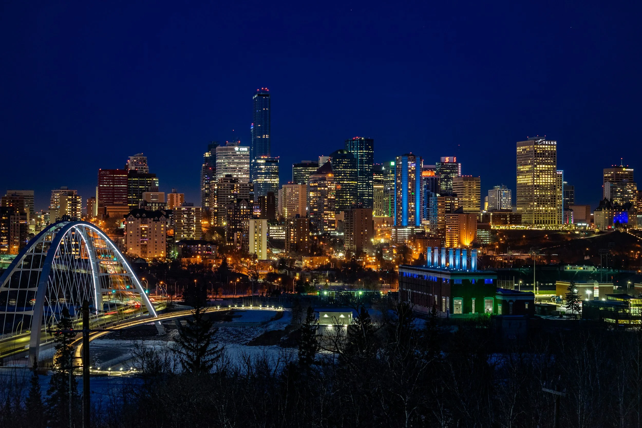 Nighttime city skyline of downtown with illuminated buildings, a busier bridge in the foreground, and some snow-covered ground in the lower part of the image.