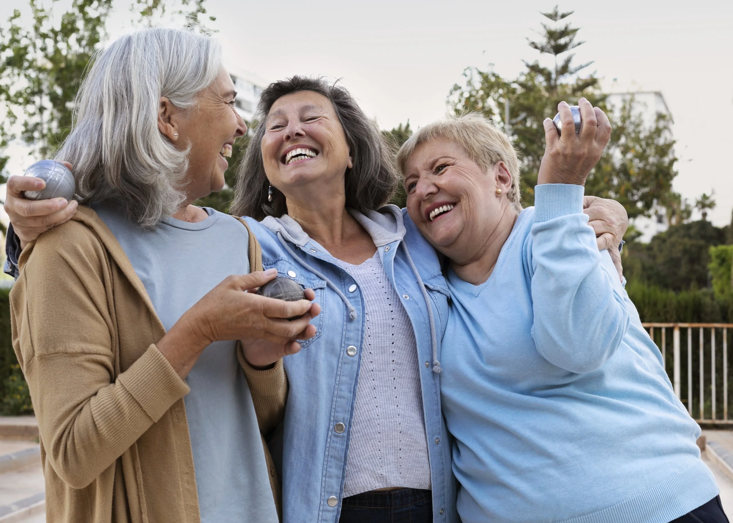 Three elderly women smiling, hugging, and holding pet rocks outdoors.