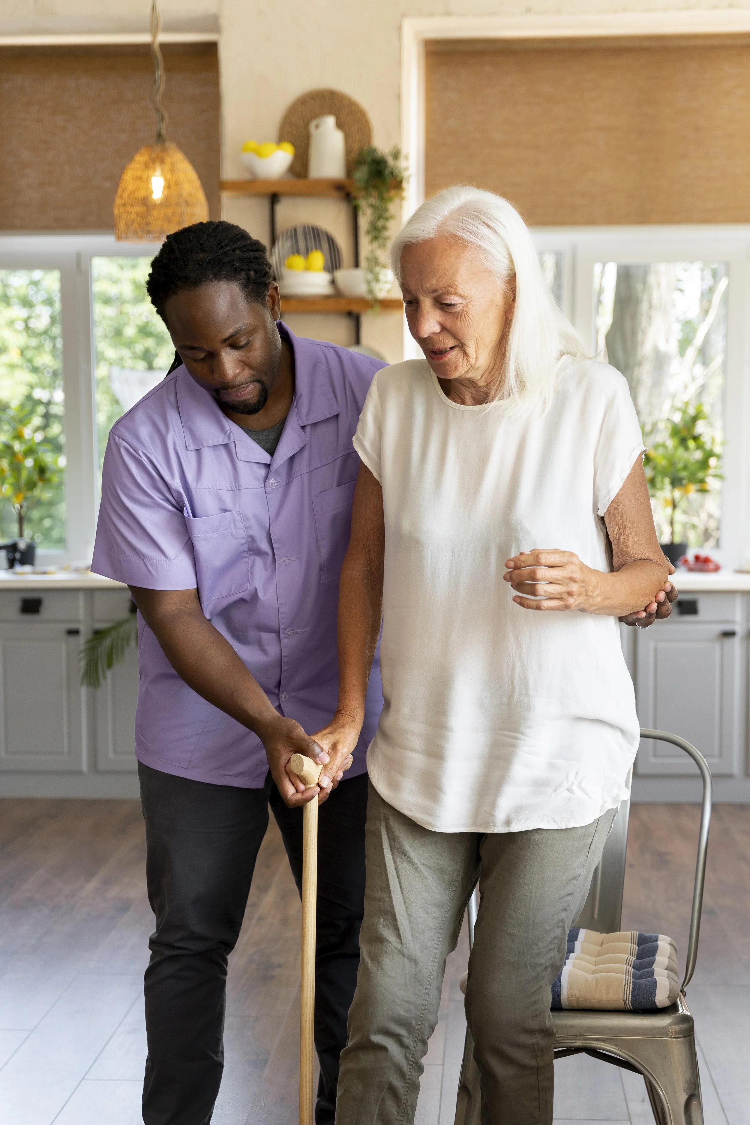A caregiver assisting an elderly woman to walk using a cane in a bright kitchen.