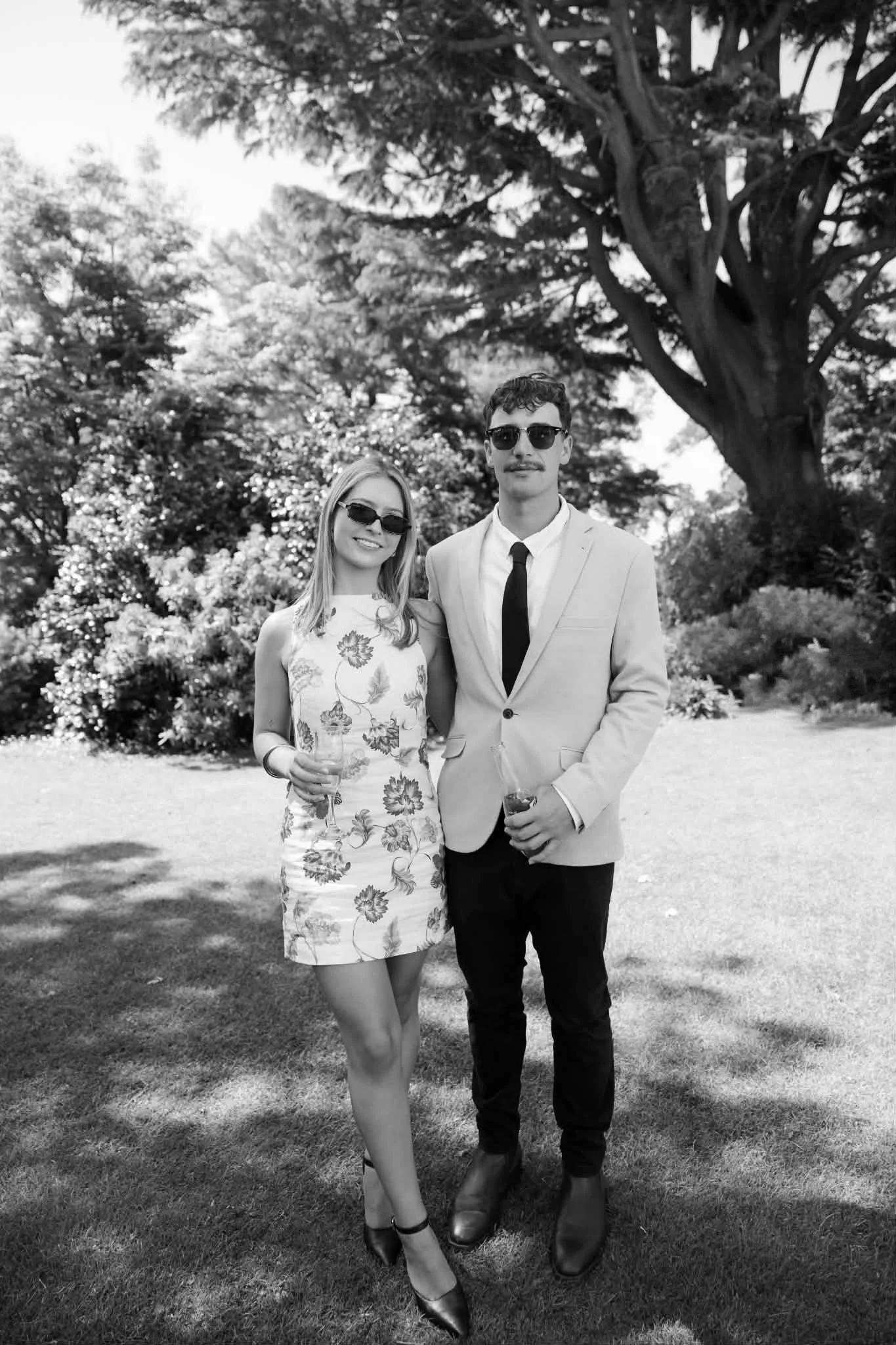 A black-and-white photo of a young woman and man standing outdoors. The woman is wearing a floral dress and sunglasses, holding a drink. The man is wearing a light-colored suit, tie, and sunglasses, also holding a drink. They are smiling with trees and bushes in the background.