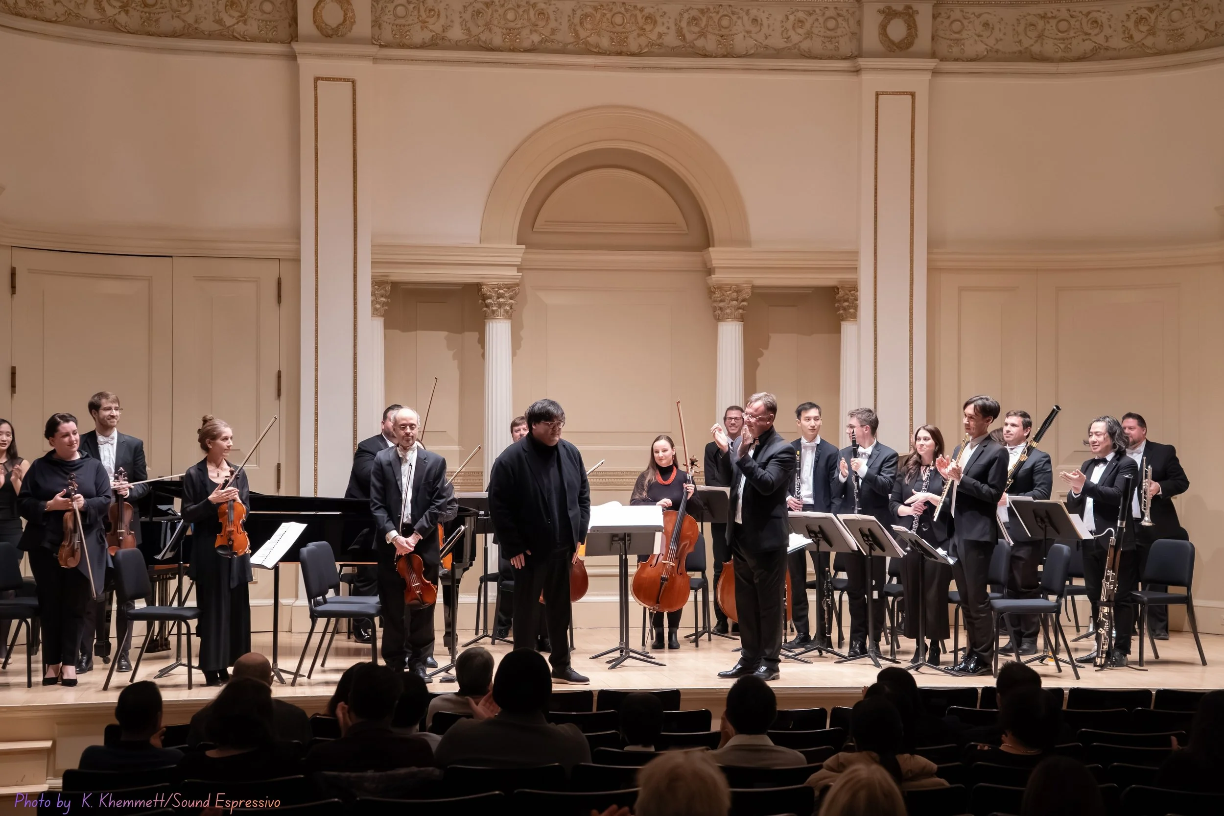 Orchestra on stage with conductors, audience in front, in a concert hall with classical architecture.
