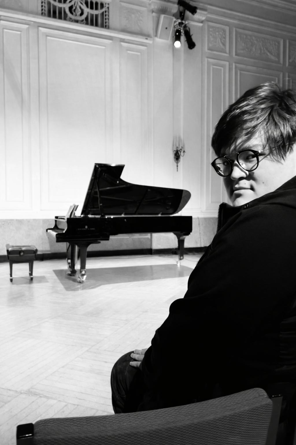 A person with glasses sitting in front of a grand piano in a music hall or concert room.