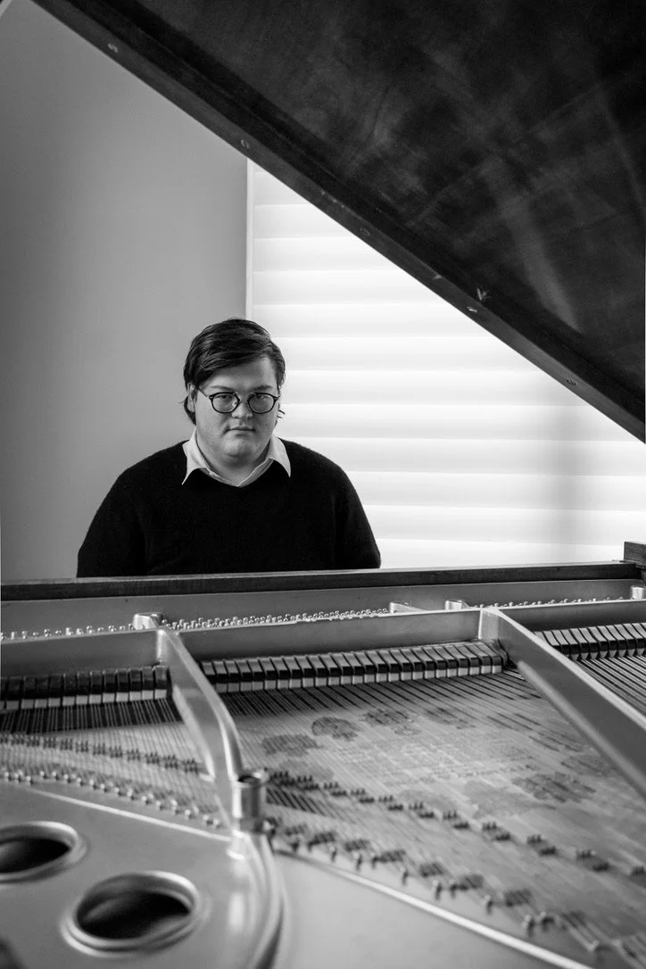 A young man with glasses and dark hair looks at the camera while sitting behind an open grand piano with the lid raised, in a room with a window covered by horizontal blinds.