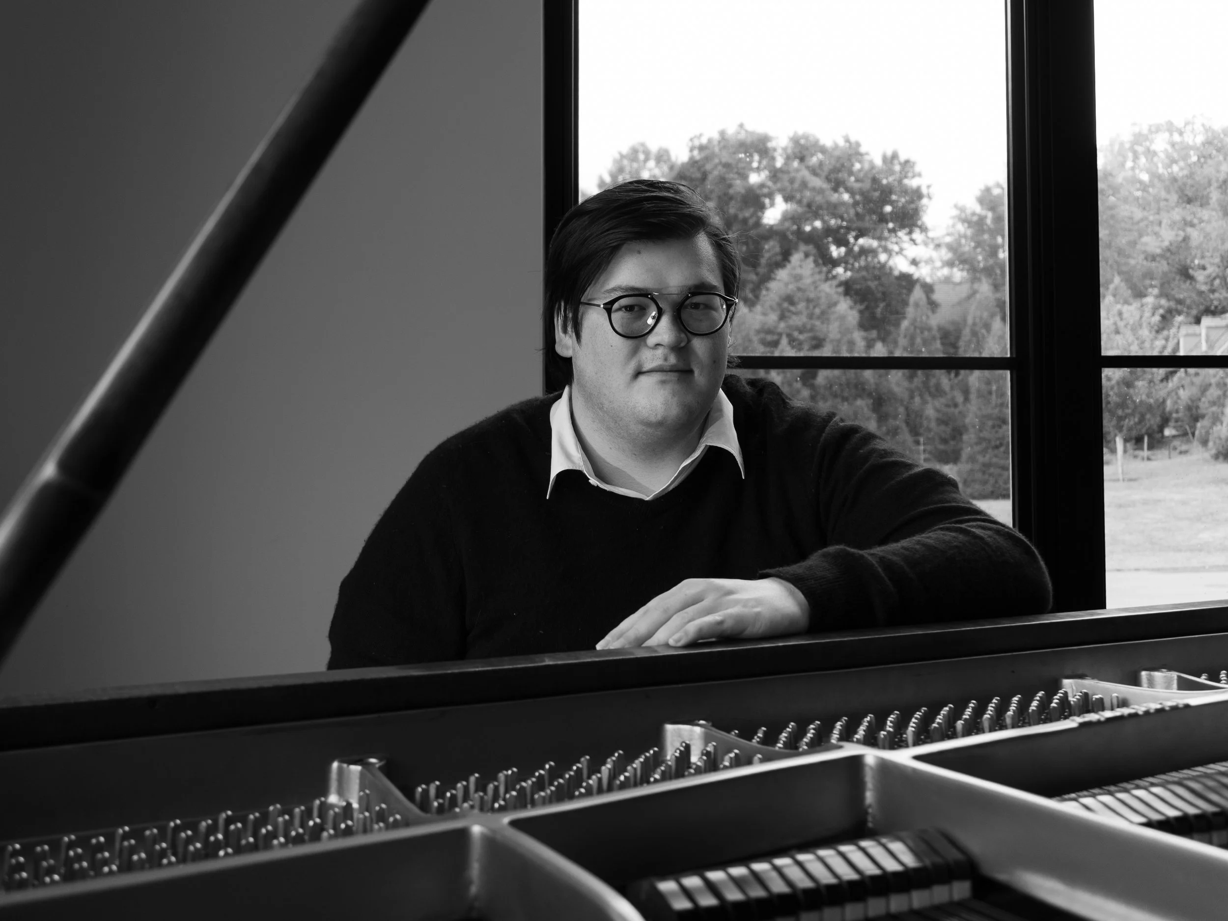 Black and white photo of a young man with glasses sitting at a grand piano, looking at the camera, with a large window and trees outside in the background.