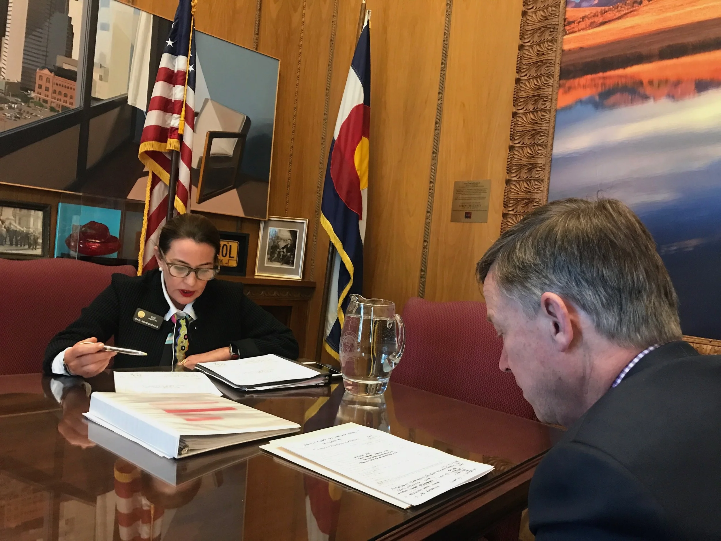 A woman in a black blazer with glasses, sitting at a table with documents, talking to a man whose head is down. The room has wooden walls, framed photos and flags of the United States and Colorado in the background.