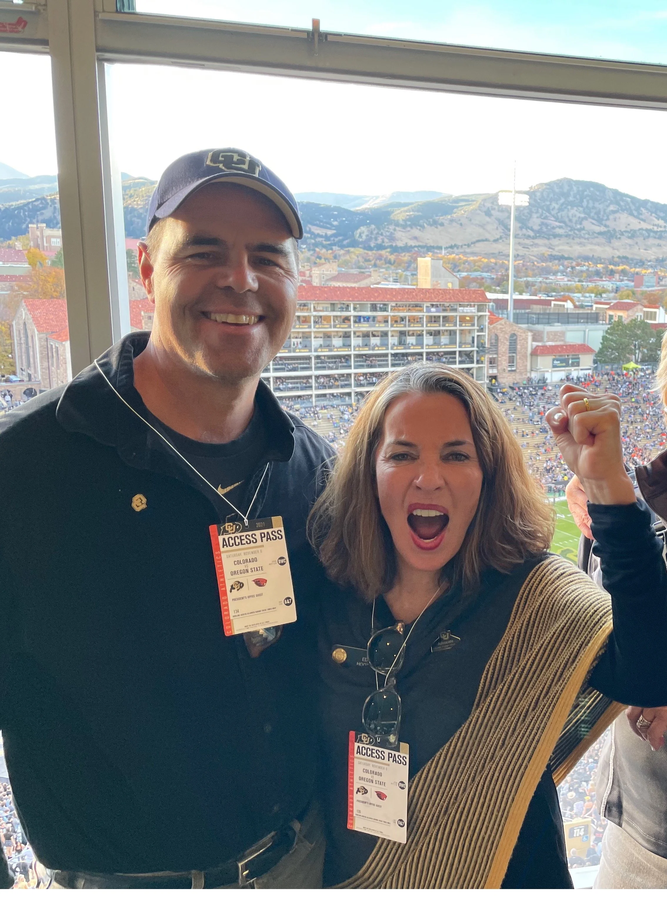 Two people at a sports stadium, the man is wearing an Oregon State cap and jacket, and the woman is flexing her arm. They are both wearing access passes, with a view of a stadium and mountains in the background.