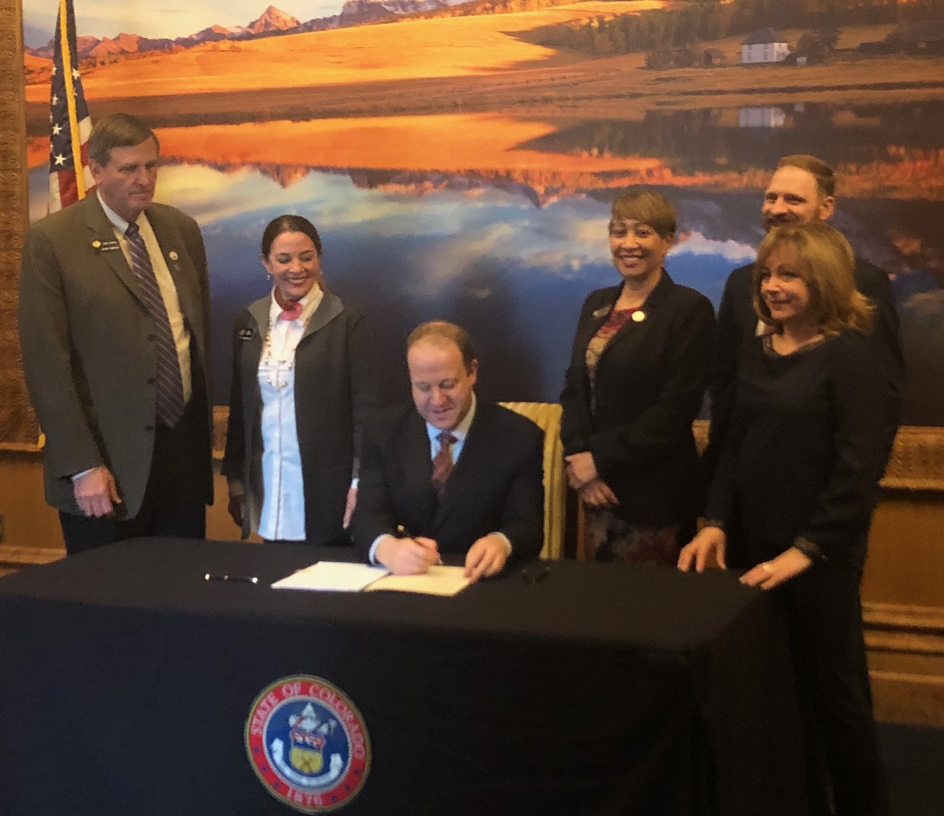 A man in a suit is seated at a table, signing a document, surrounded by five others standing behind him. The backdrop features a large landscape painting of a lake and mountains at sunset. The table has a Colorado state emblem on the front.