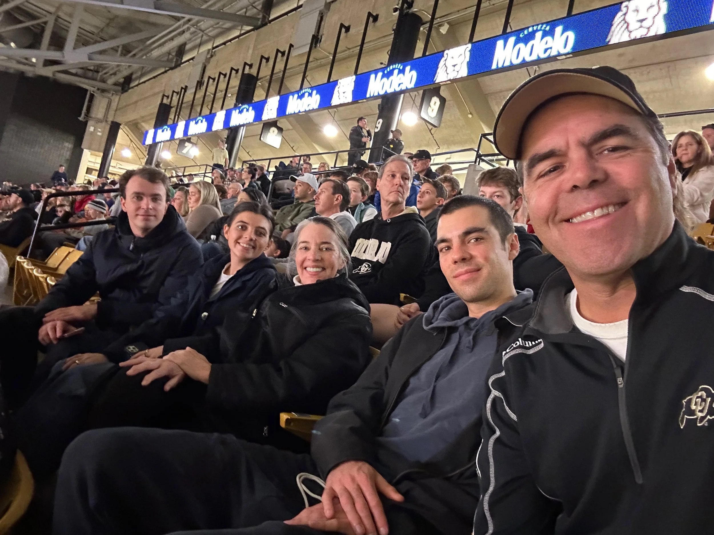 People sitting in stadium seats at a sporting event, smiling and looking at the camera, with a large digital advertisement board overhead.