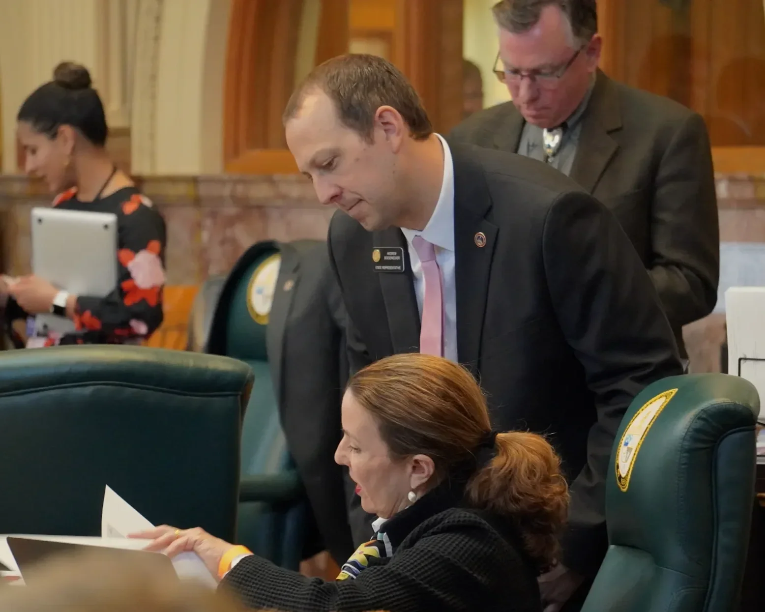 A man in a dark suit leaning over a woman seated at a desk, holding papers, with a person in the background looking at a tablet in a formal setting.