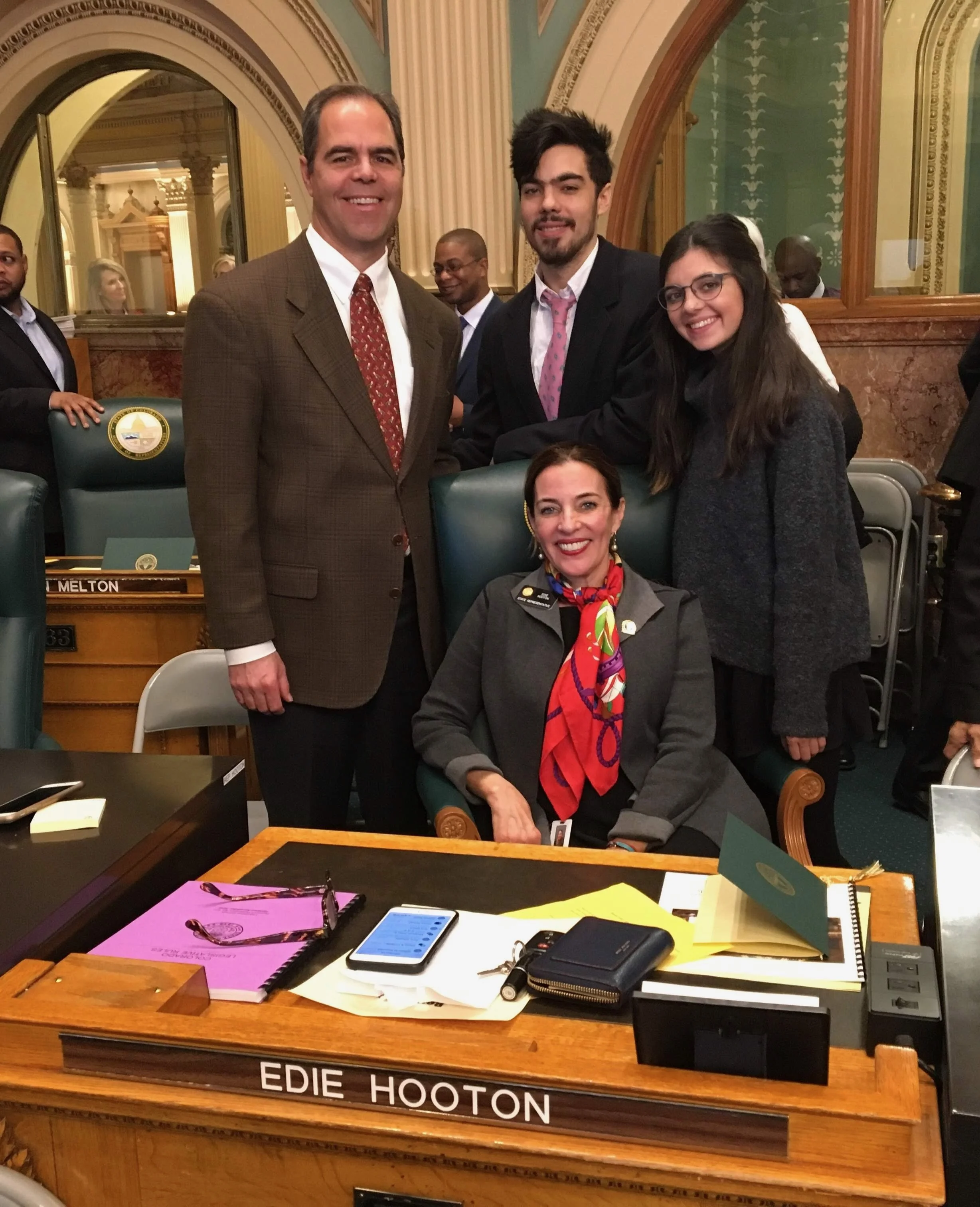 A woman sitting at a desk with a nameplate reading 'Edie Hooton' is surrounded by four people standing, all smiling and posing for the photo in a formal setting with ornate decor.