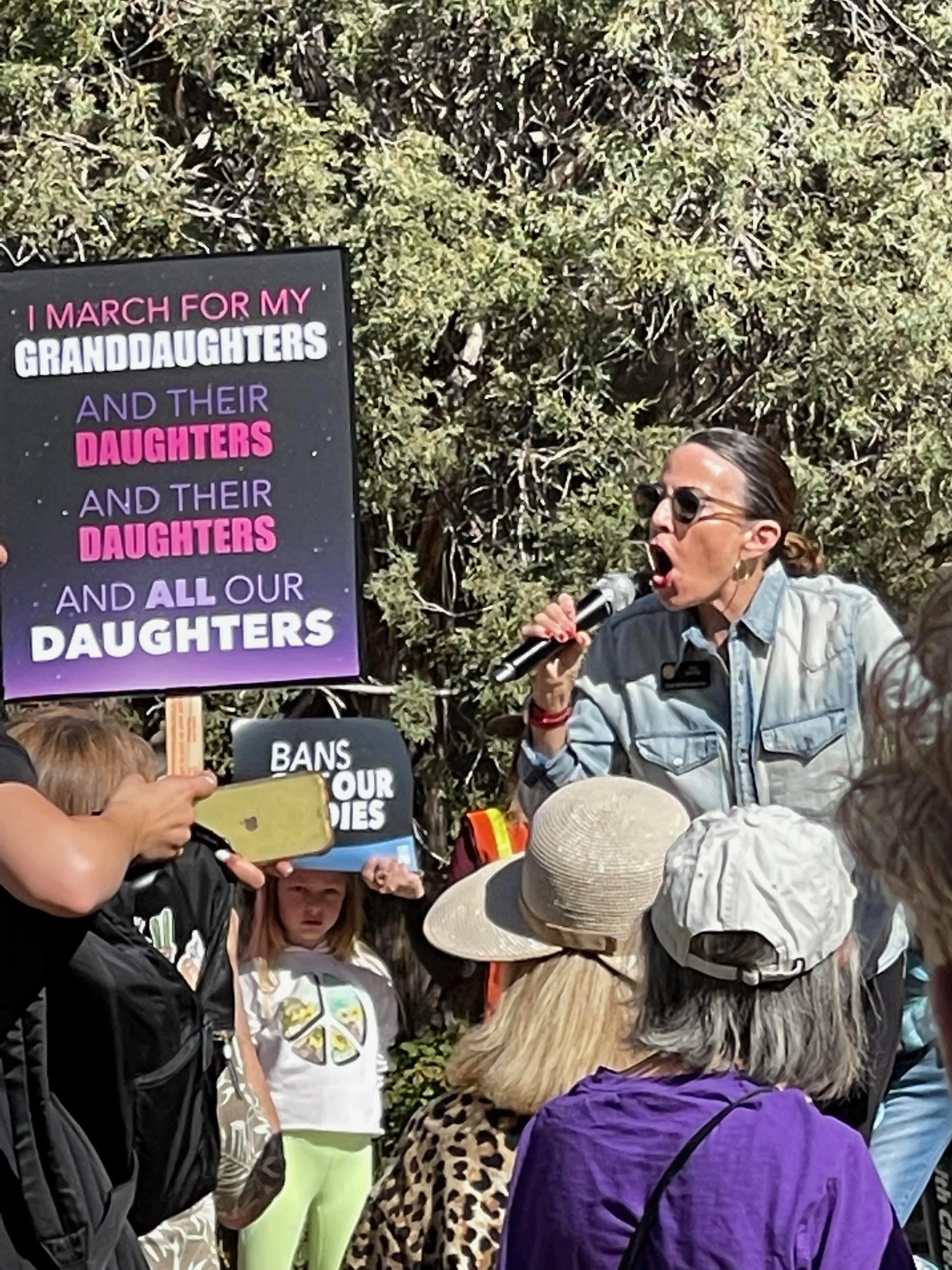 A woman speaking into a microphone at a protest or rally, with a sign that reads "I march for my granddaughters and their daughters and their daughters and all our daughters". Several people are gathered around, some holding signs and wearing hats.