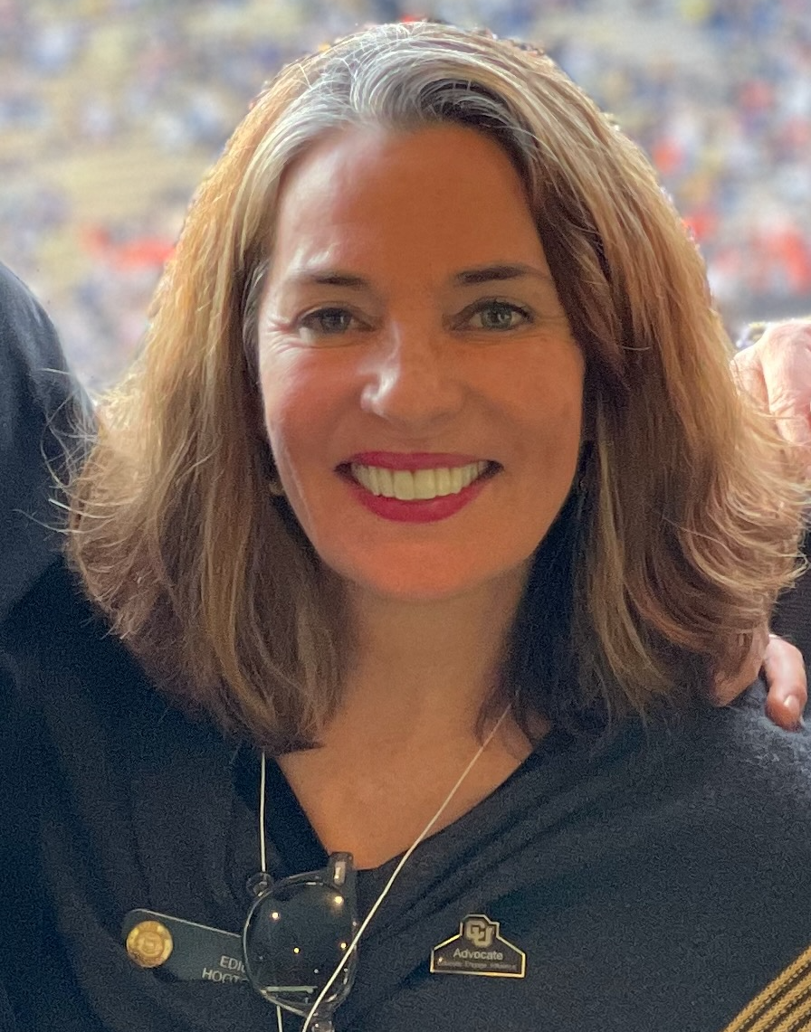 A woman with shoulder-length light brown hair and red lipstick, smiling, wearing a dark top with a name badge and advocate pin, with an outdoor or stadium background.