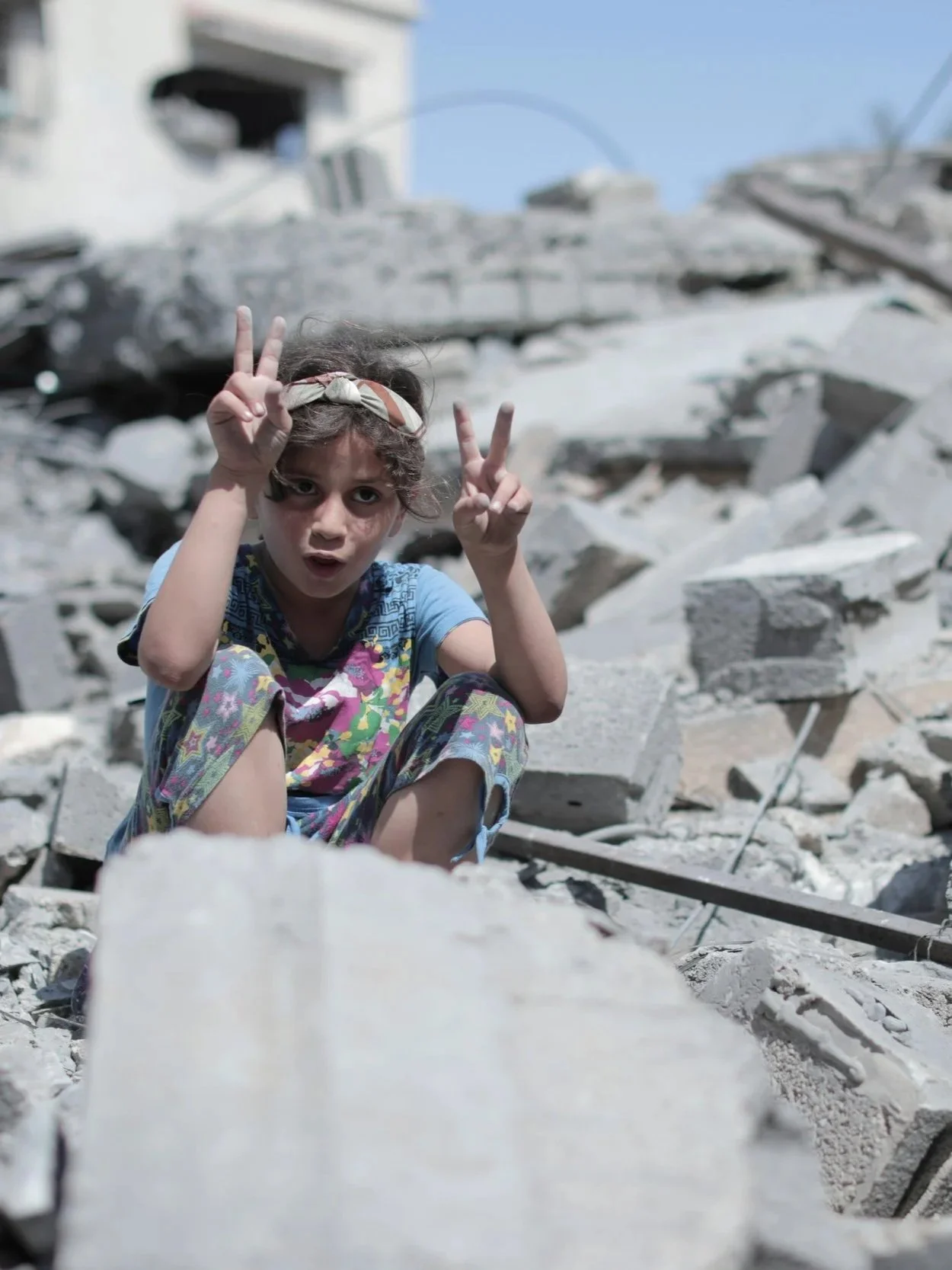A young girl sitting among rubble and debris after a building collapse, making peace signs with both hands.
