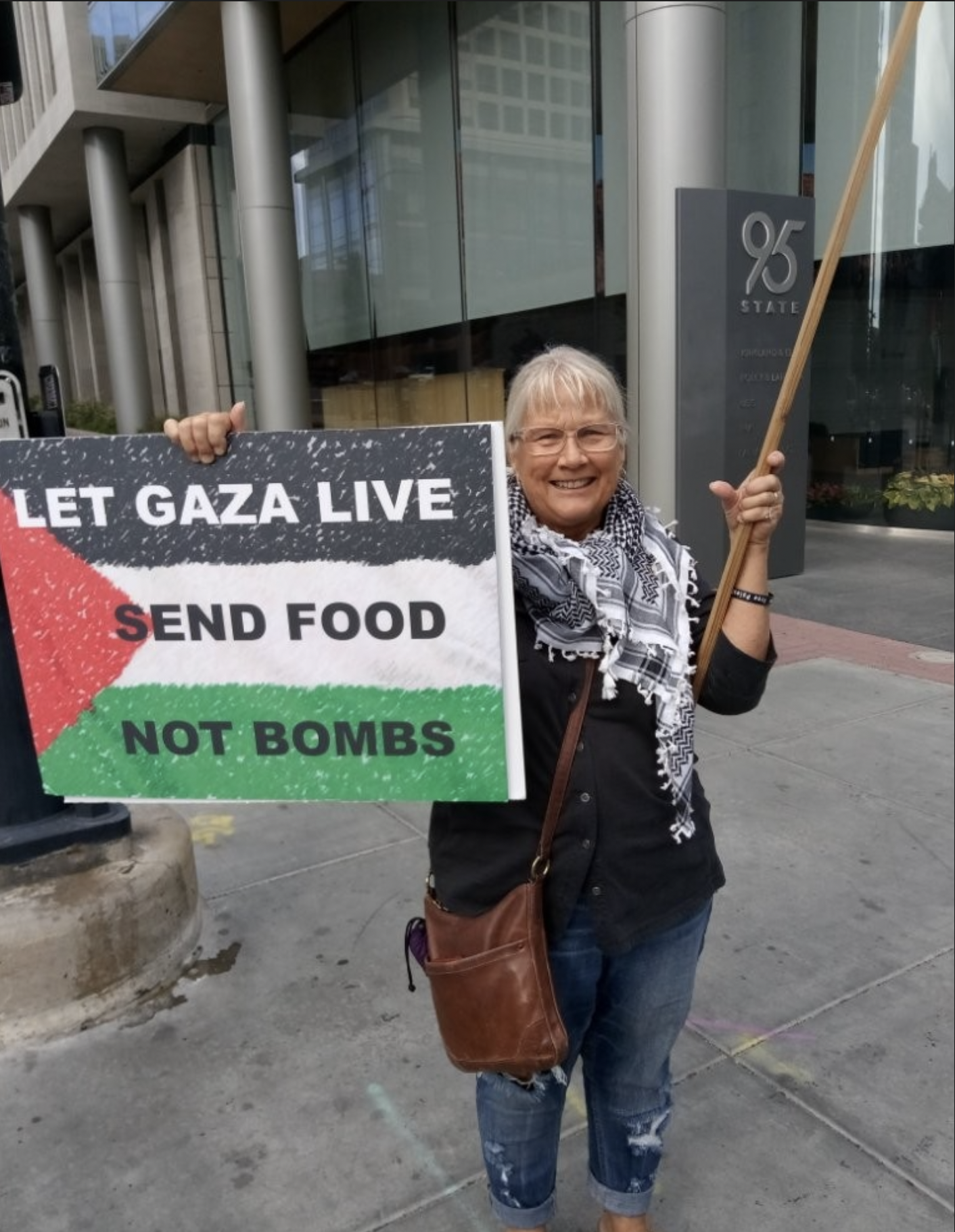 A woman standing on a sidewalk holding a sign that says "Let Gaza Live, Send Food, Not Bombs" with the Palestinian flag colors. She is smiling and wearing glasses, a black shirt, a black-and-white scarf, and has a brown shoulder bag. Behind her is a modern building entrance with the number 95 on a sign.