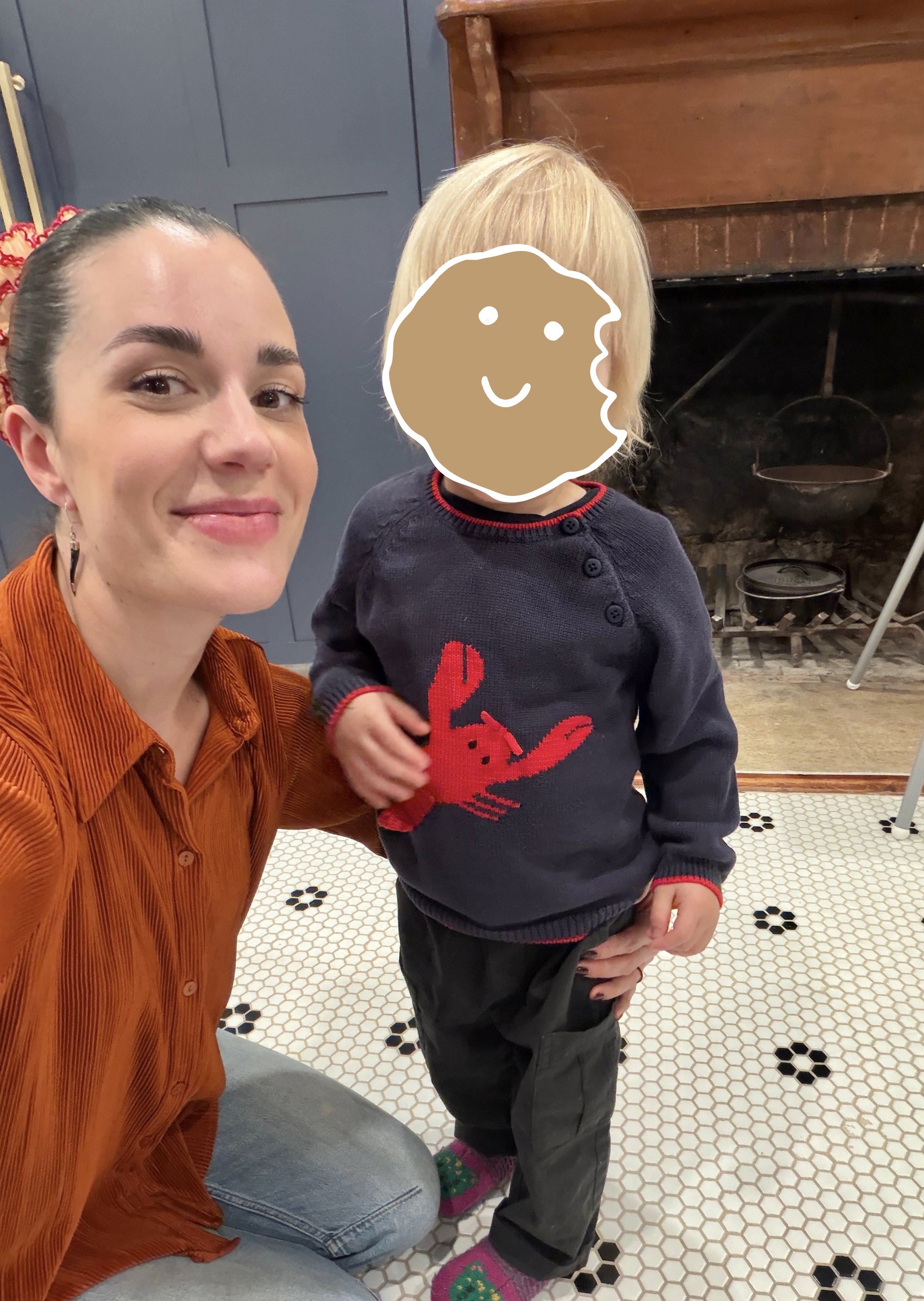 A woman kneeling with a small blond child on a tiled kitchen floor, the child's face is covered by a cookie