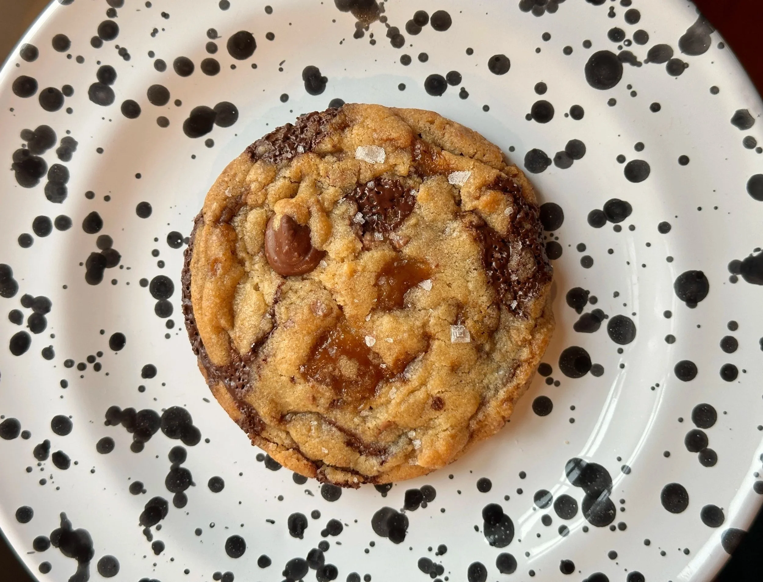 A chocolate chip cookie on a white plate with black splatter patterns.