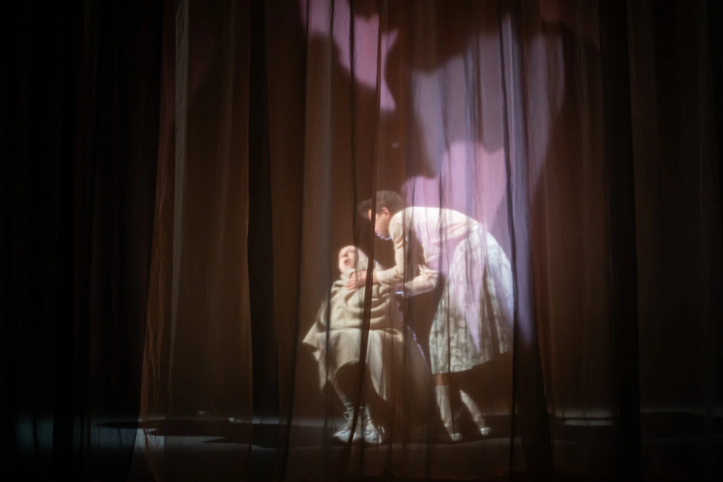 A woman and child behind stage curtains, with the woman bending down to help the child, illuminated by stage lights.