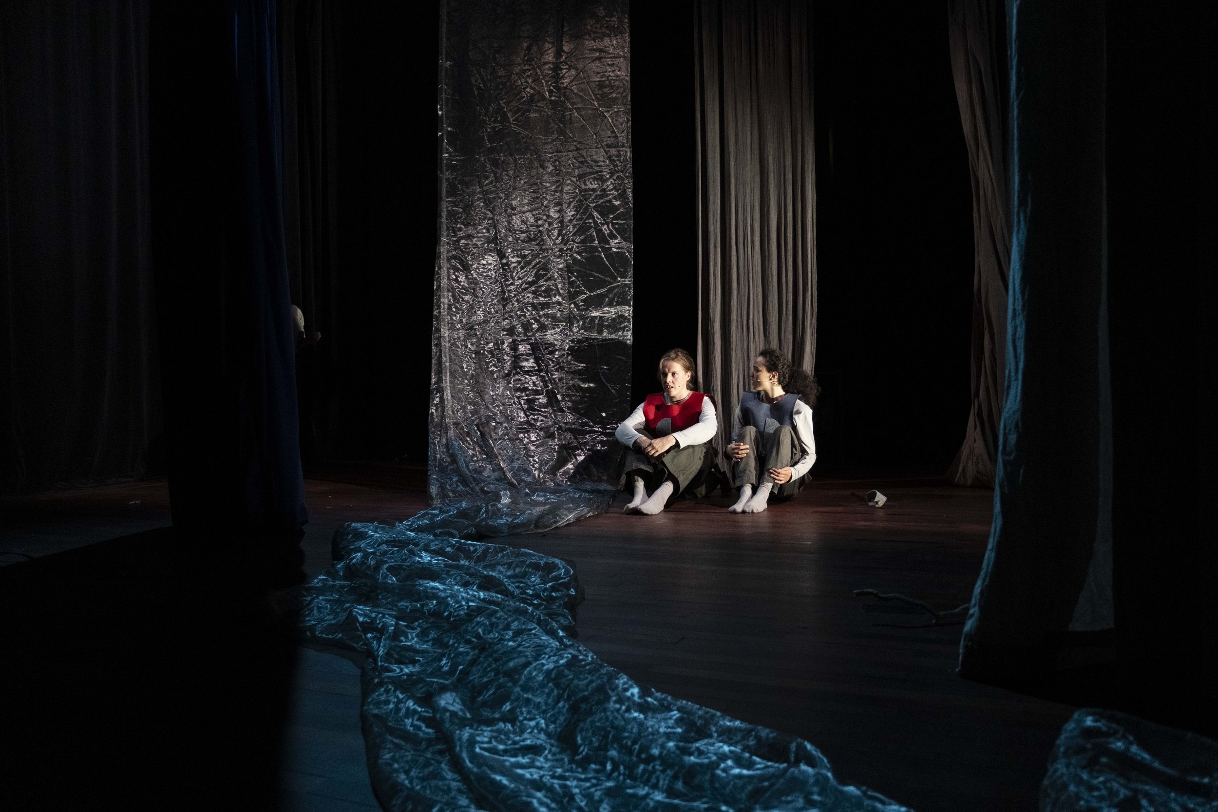 Two children sitting on the stage floor, appearing to be involved in a theatrical performance, with dark curtains behind them and crumpled plastic sheets on the stage.