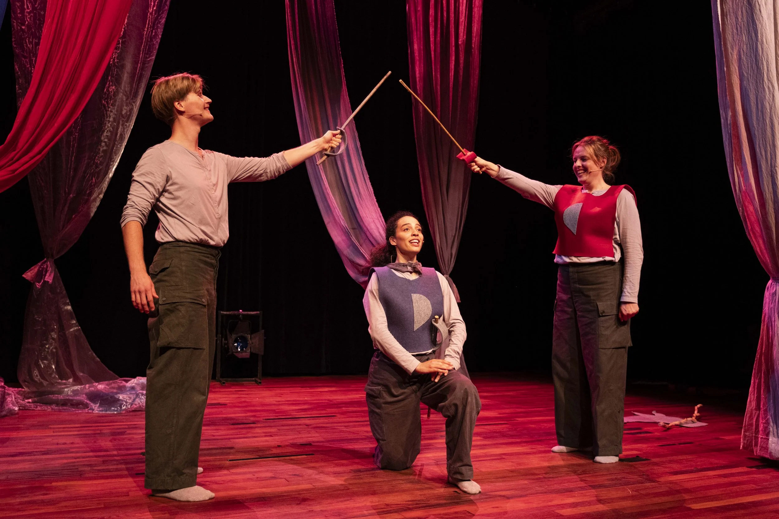 Three women on stage performing a play with a sword fight scene. Two women standing, holding swords, and one kneeling with hands clasped, all with theatrical costumes and colorful curtains in the background.