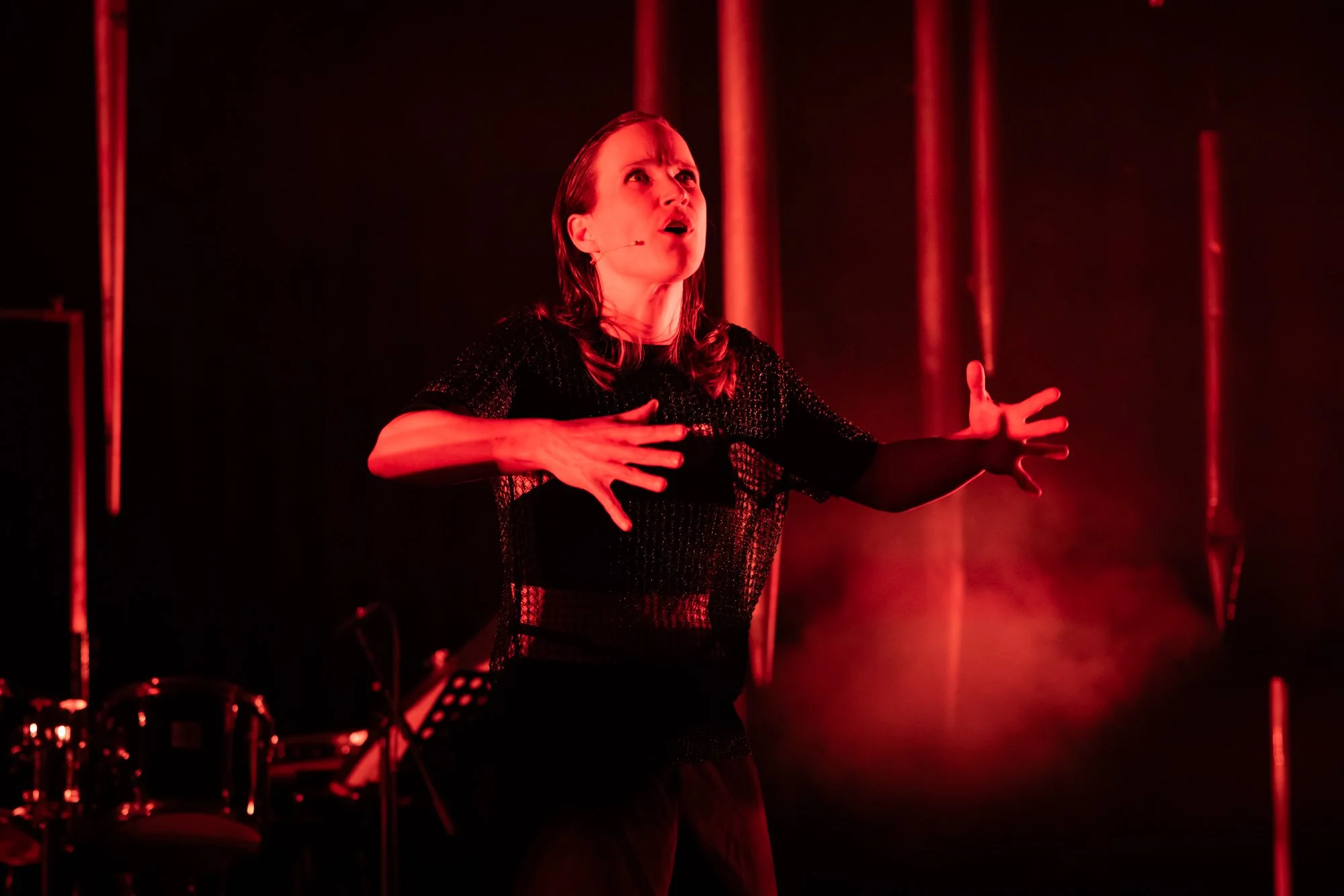 A woman passionately singing or acting on stage, with red lighting and curtains in the background, wearing black attire.