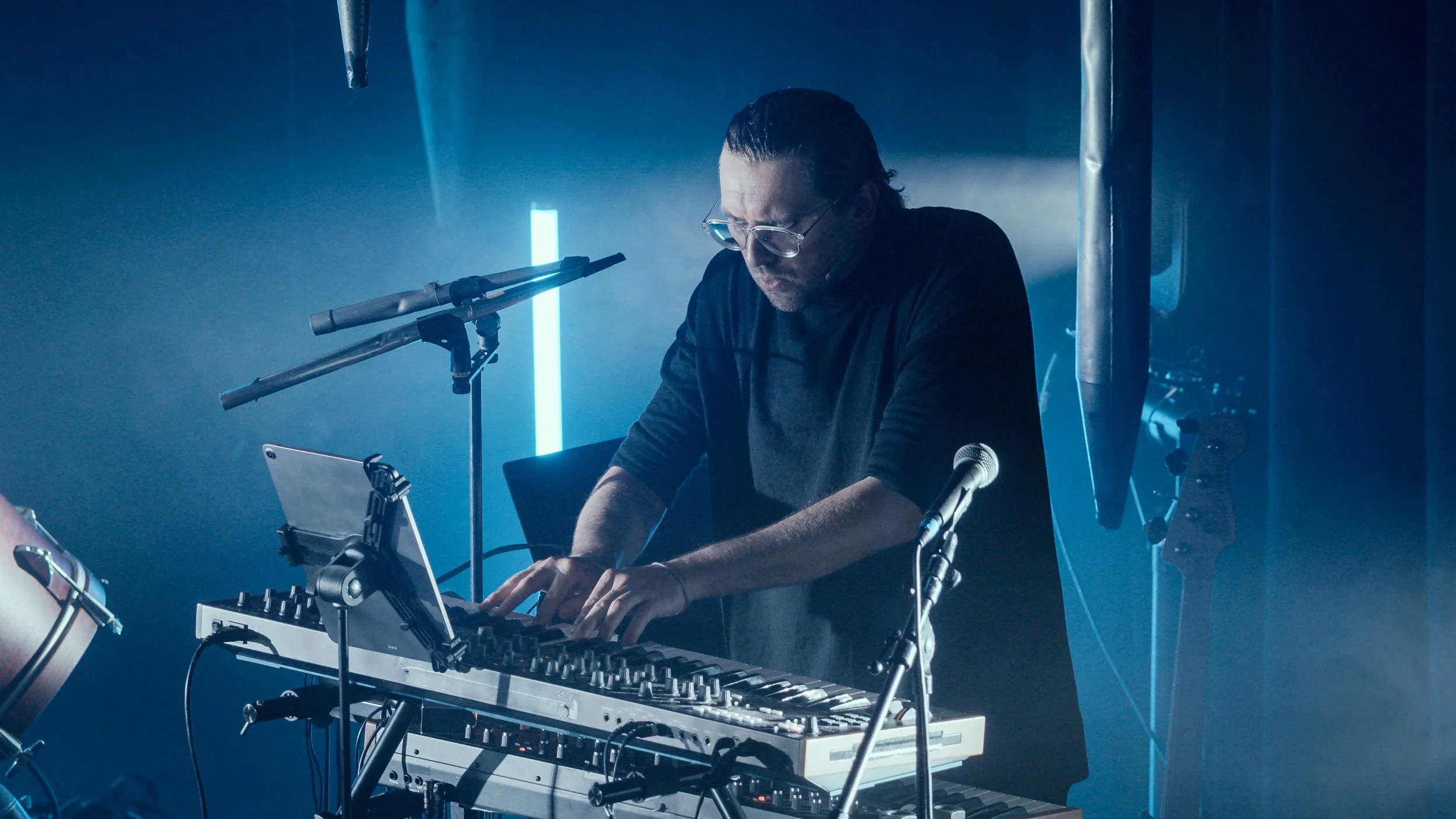 A man with glasses and slicked-back hair operating a music keyboard on stage, surrounded by microphones, with blue lighting and smoke in the background.