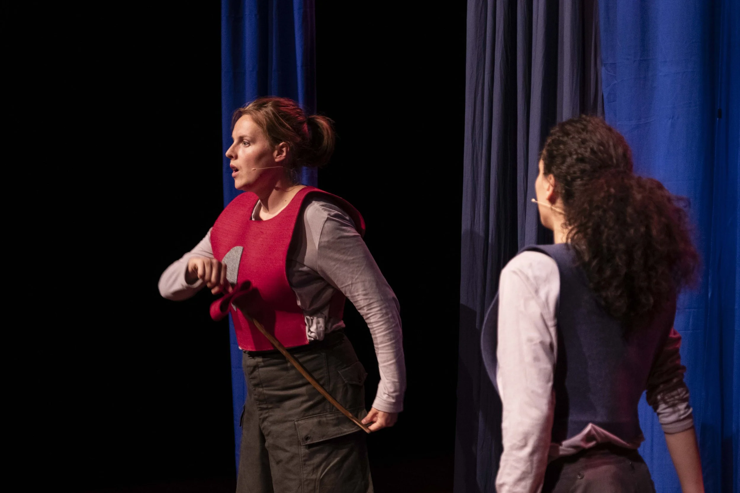 Two women on stage during a theatrical performance, one dressed as a lion with a red mane and the other as a woman with curly hair, in front of blue curtains.