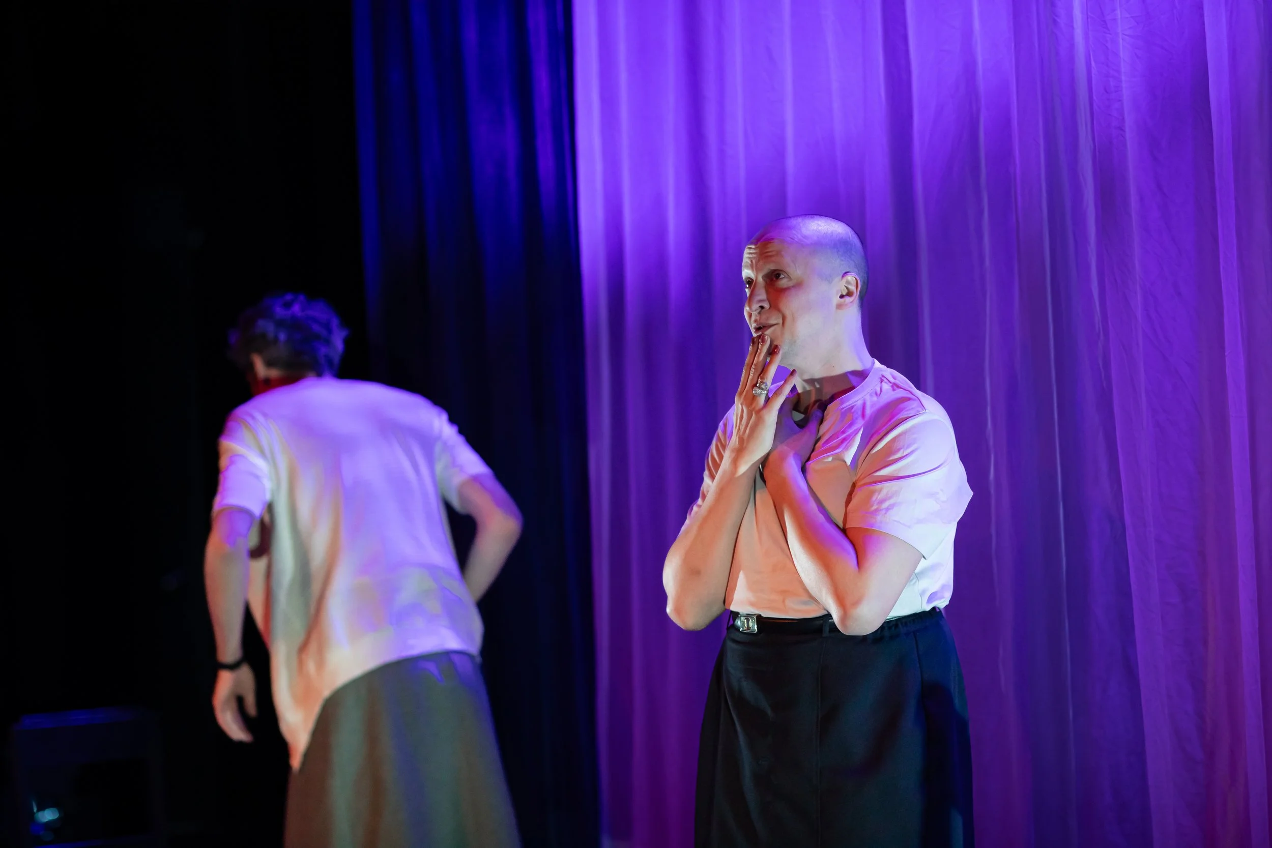 Two performers on stage under purple lighting, one with a shaved head and hand on chest, the other with back turned wearing a white top and dark skirt.