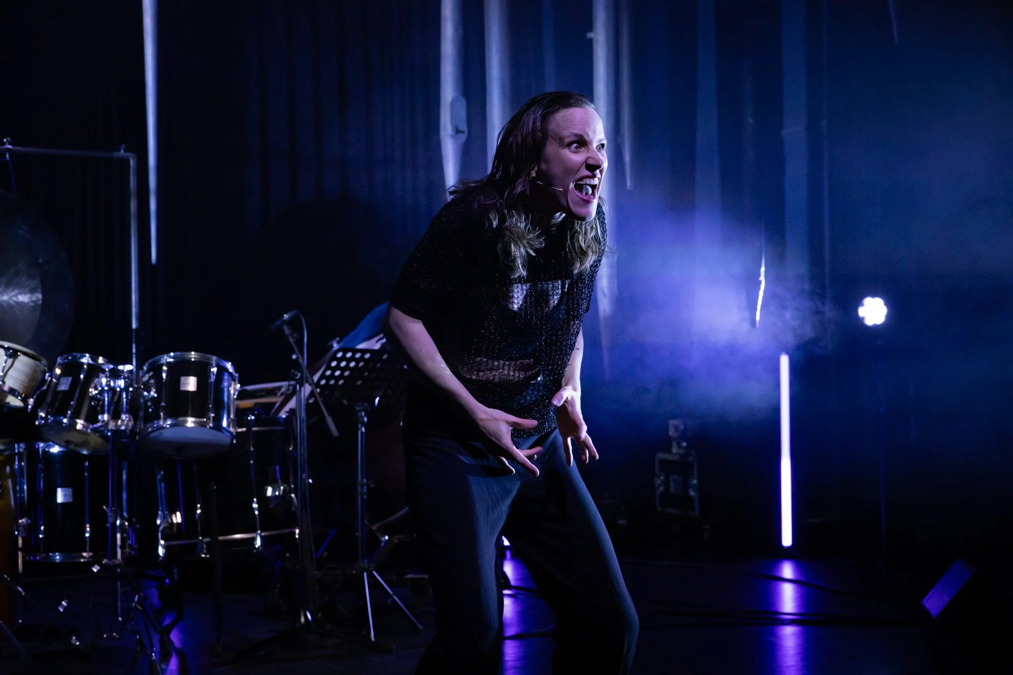 A woman on stage passionately performing, with drums and musical equipment in the background, illuminated by blue and purple stage lights.