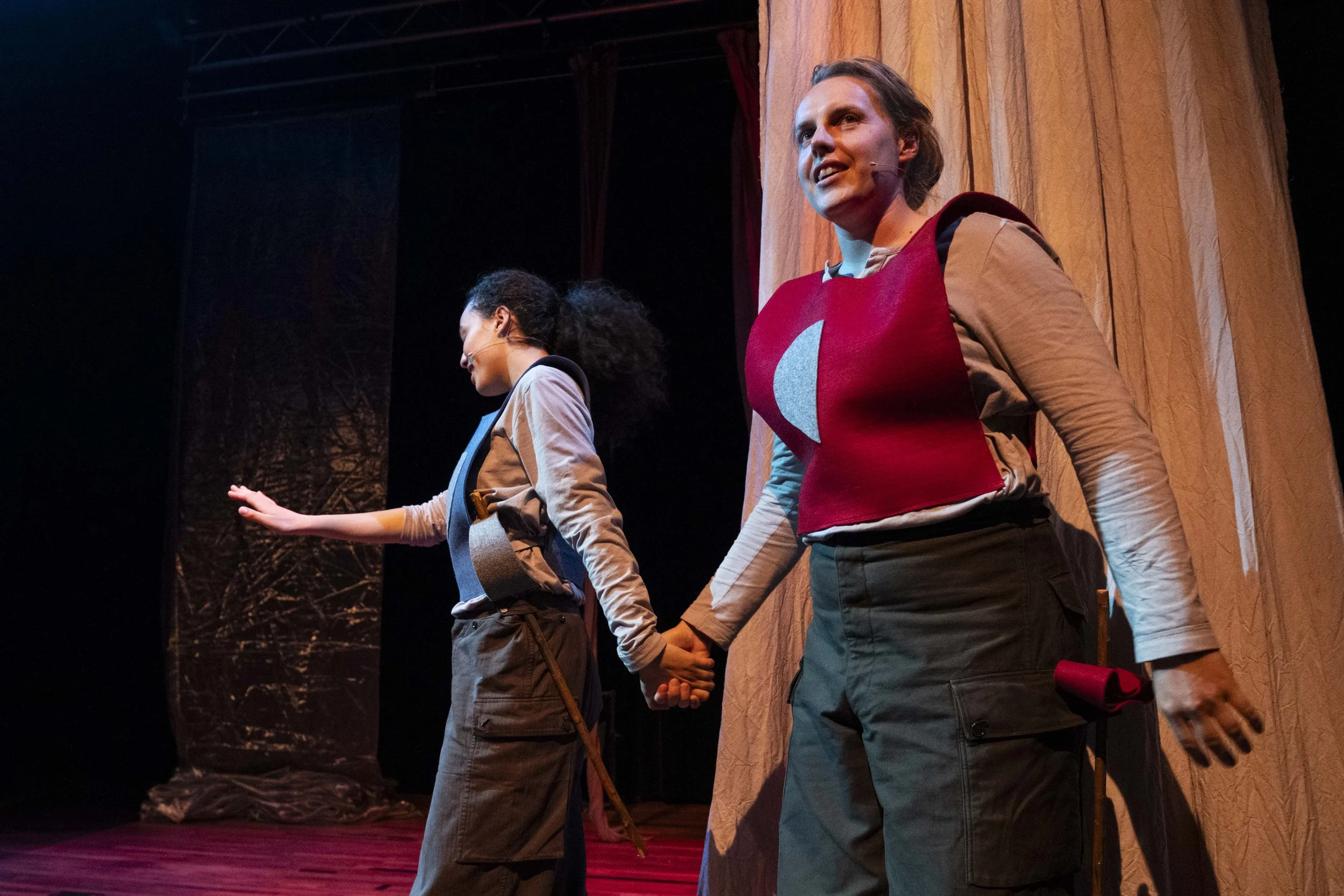 Two women dressed in costumes on a stage, holding hands with their backs to the audience, appearing to take a bow or acknowledge applause after a performance.