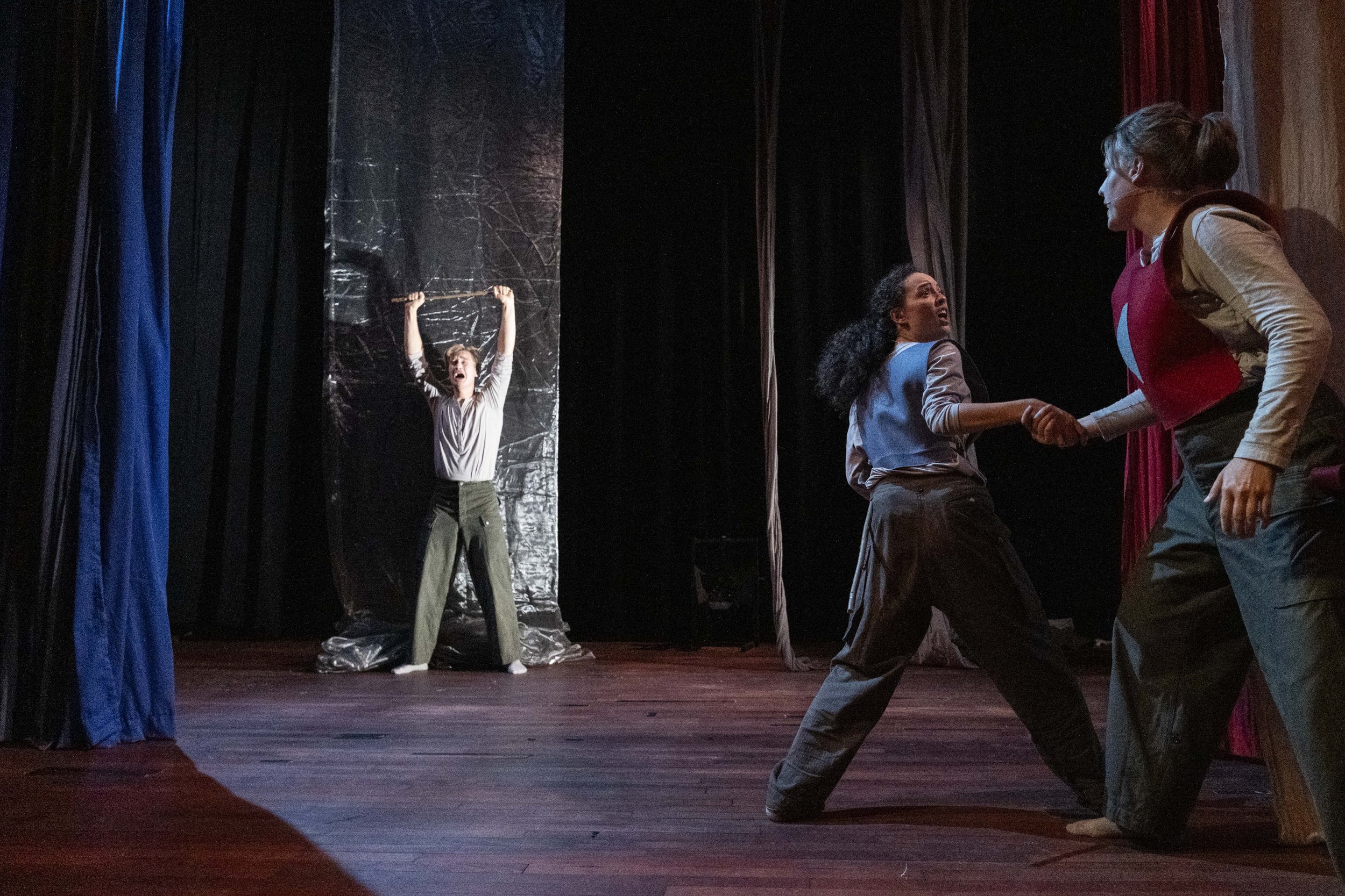 Three women acting on stage with theatrical curtains, one in the background with arms raised and distressed, and two in the foreground holding hands and engaging in an intense scene.