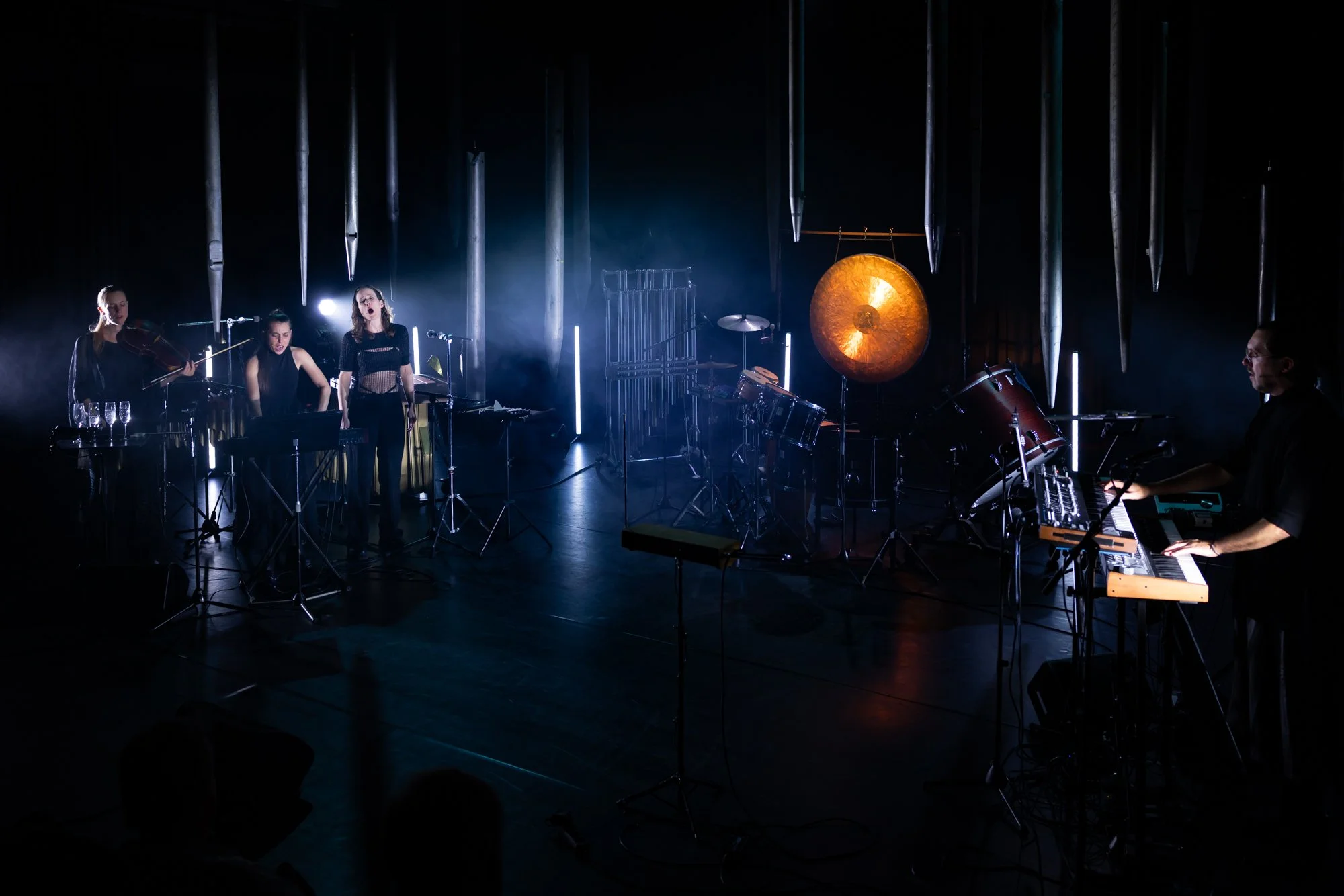 A musical performance with four female musicians on stage, surrounded by drums, percussion instruments, and a keyboard, with a large gong and metallic tubes hanging above.