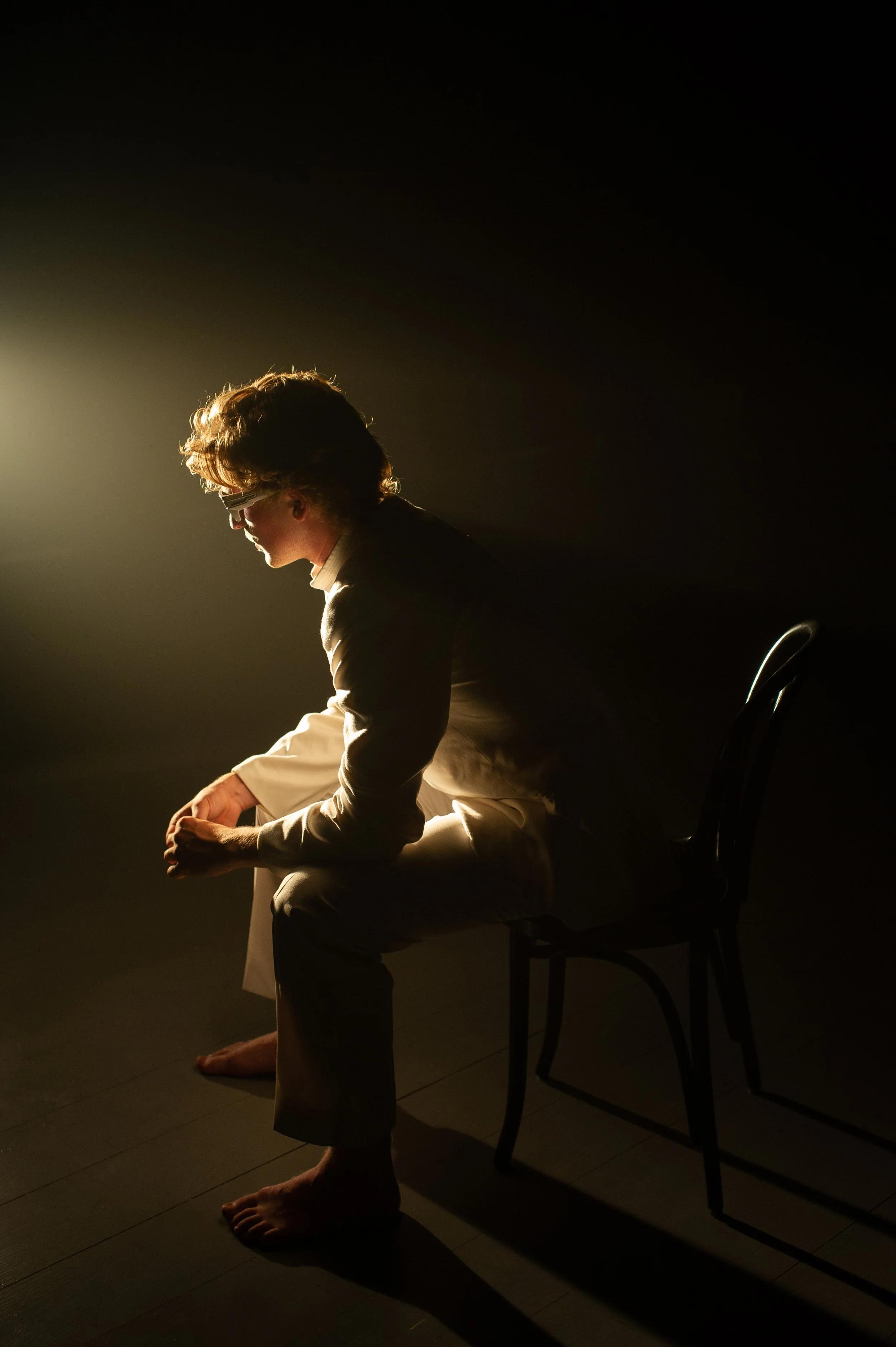 A person sitting barefoot on a chair in a dark room, illuminated from the side with a bright soft light.