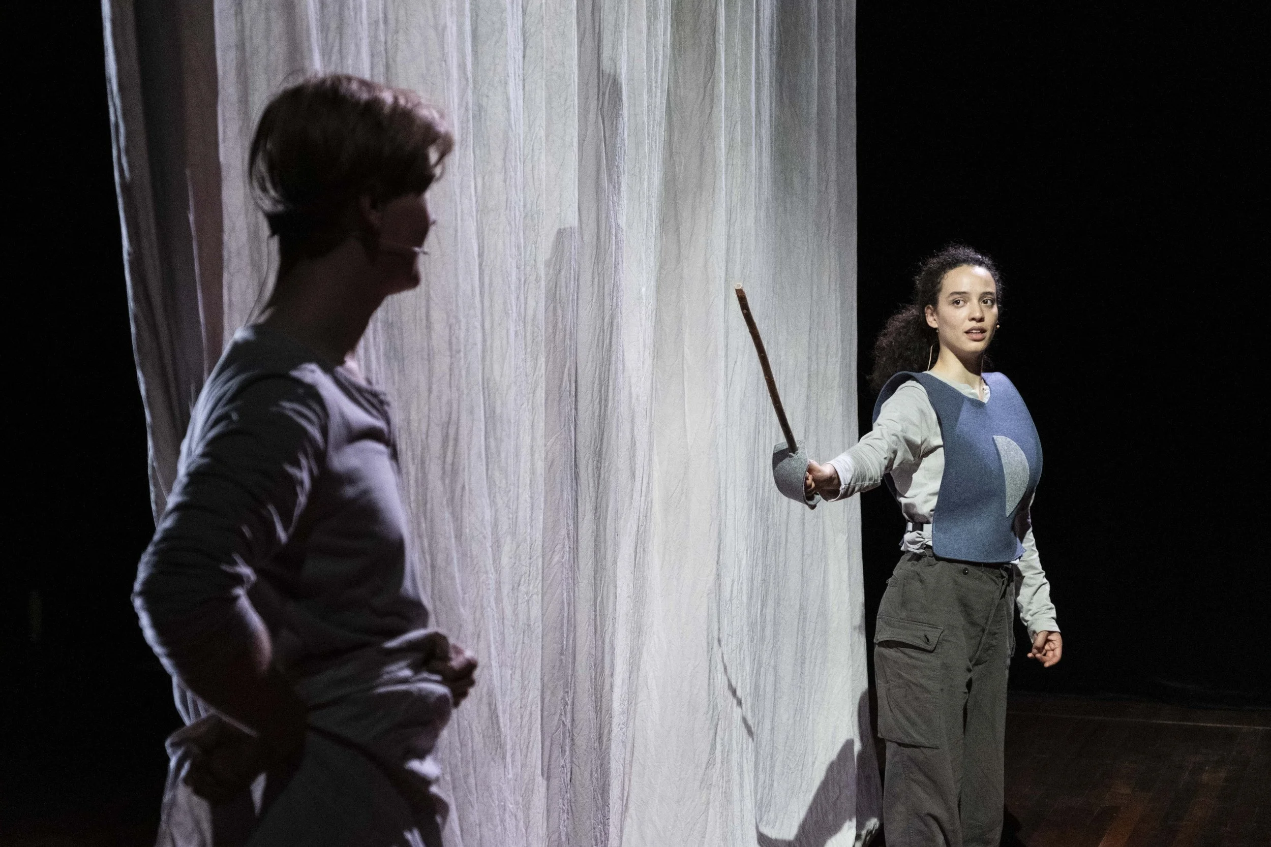 Two women backstage before a theater performance, one holding a sword, with a curtain in the background.