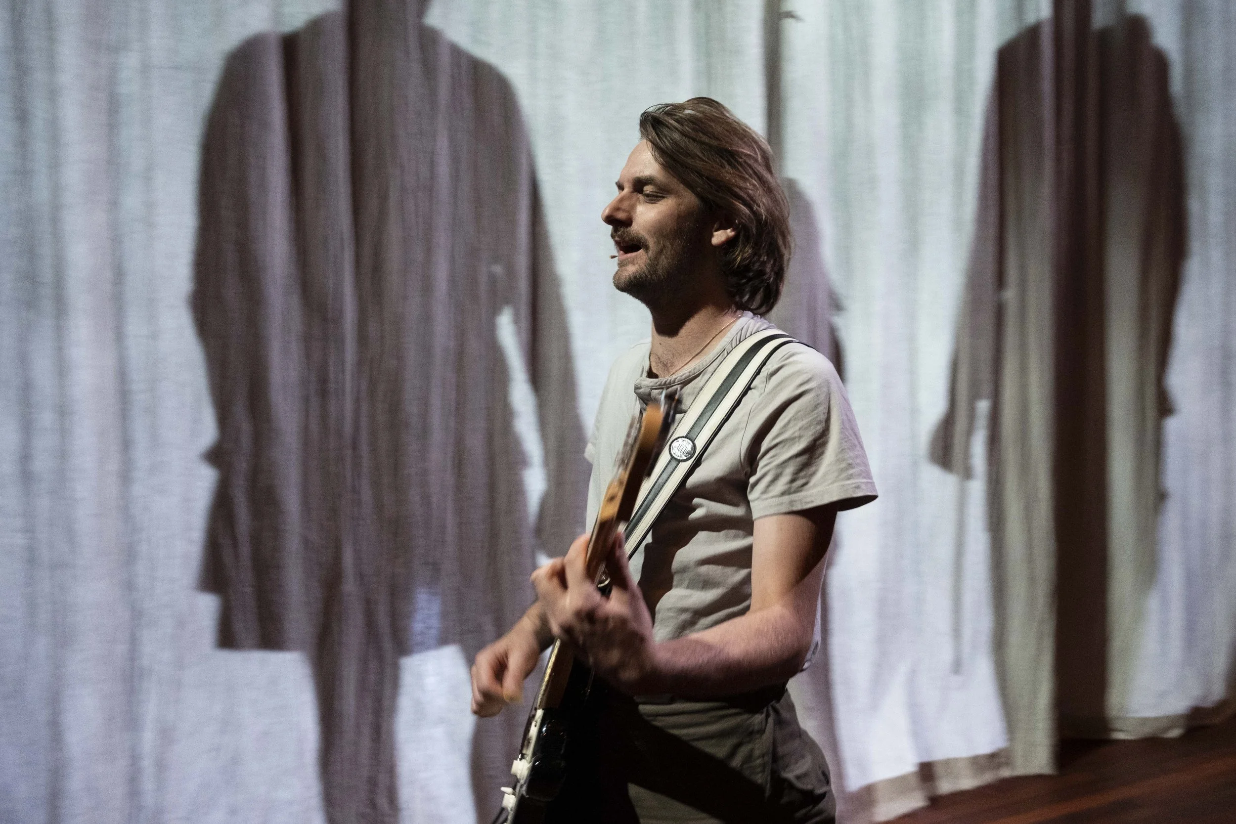 A man with long brown hair playing an electric guitar, standing in front of a backdrop with hanging shirts casting shadows.