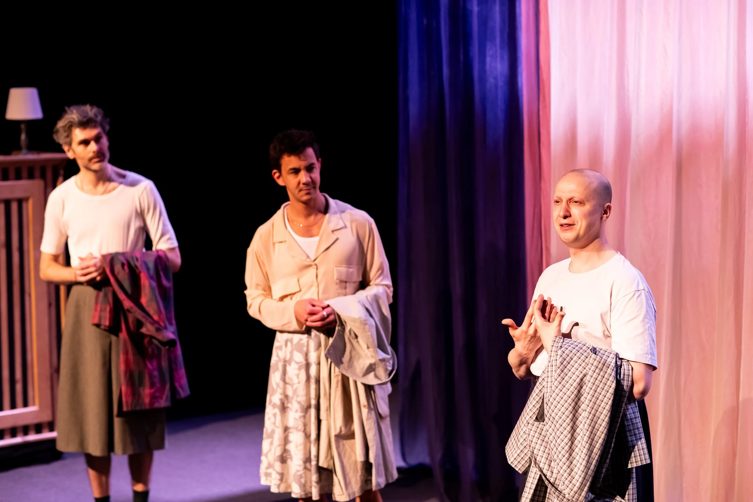 Three actors on stage performing in a play, with a woman on the right gesturing and speaking, and two women on the left listening attentively. The stage has a background with dark curtains and a wooden side table with a lamp.