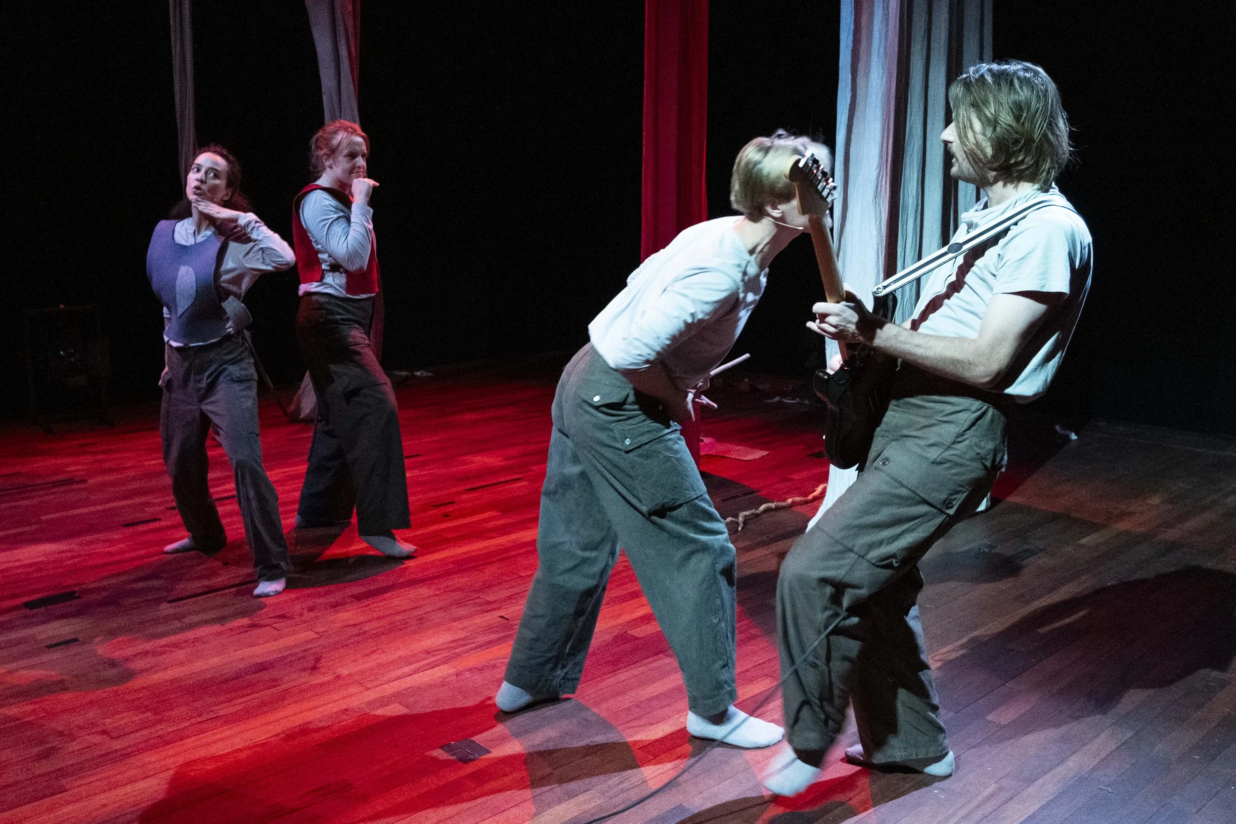 Four people rehearsing on a stage with wooden flooring, red and blue curtains, and a black background. Two women are standing in the back with their hands near their faces, while a man and a woman are at the front, with the man playing an electric gu
