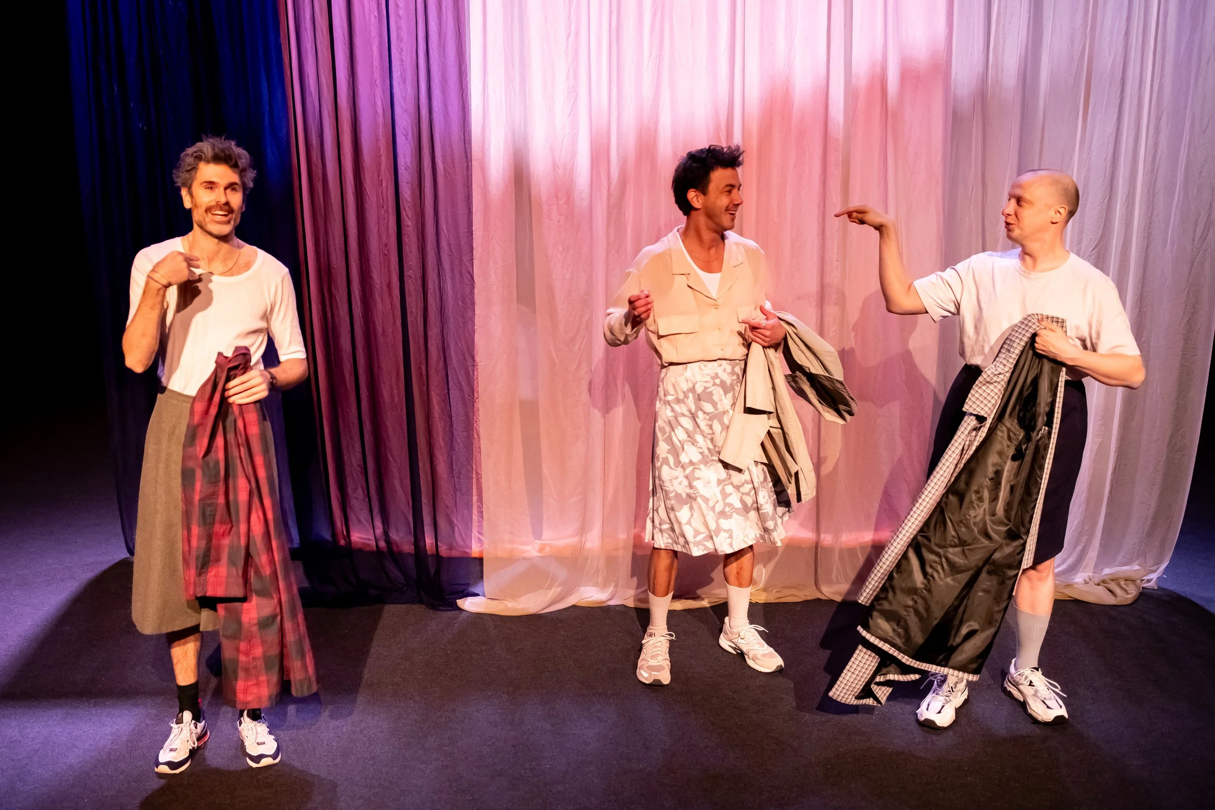 Three actors on stage in casual clothing, engaging in a comedic scene, with pink and purple curtains in the background.