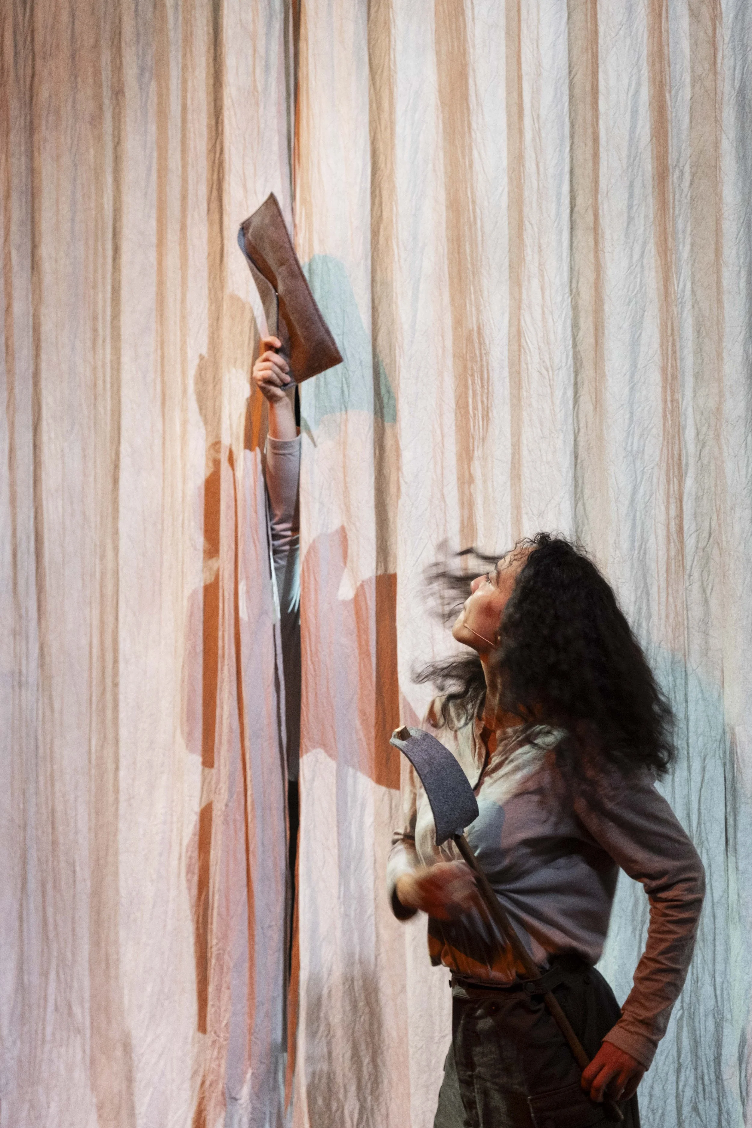 A woman with curly hair holding an axe, smiling, and engaging with a clump of hay in front of a textured, multicolored fabric backdrop.