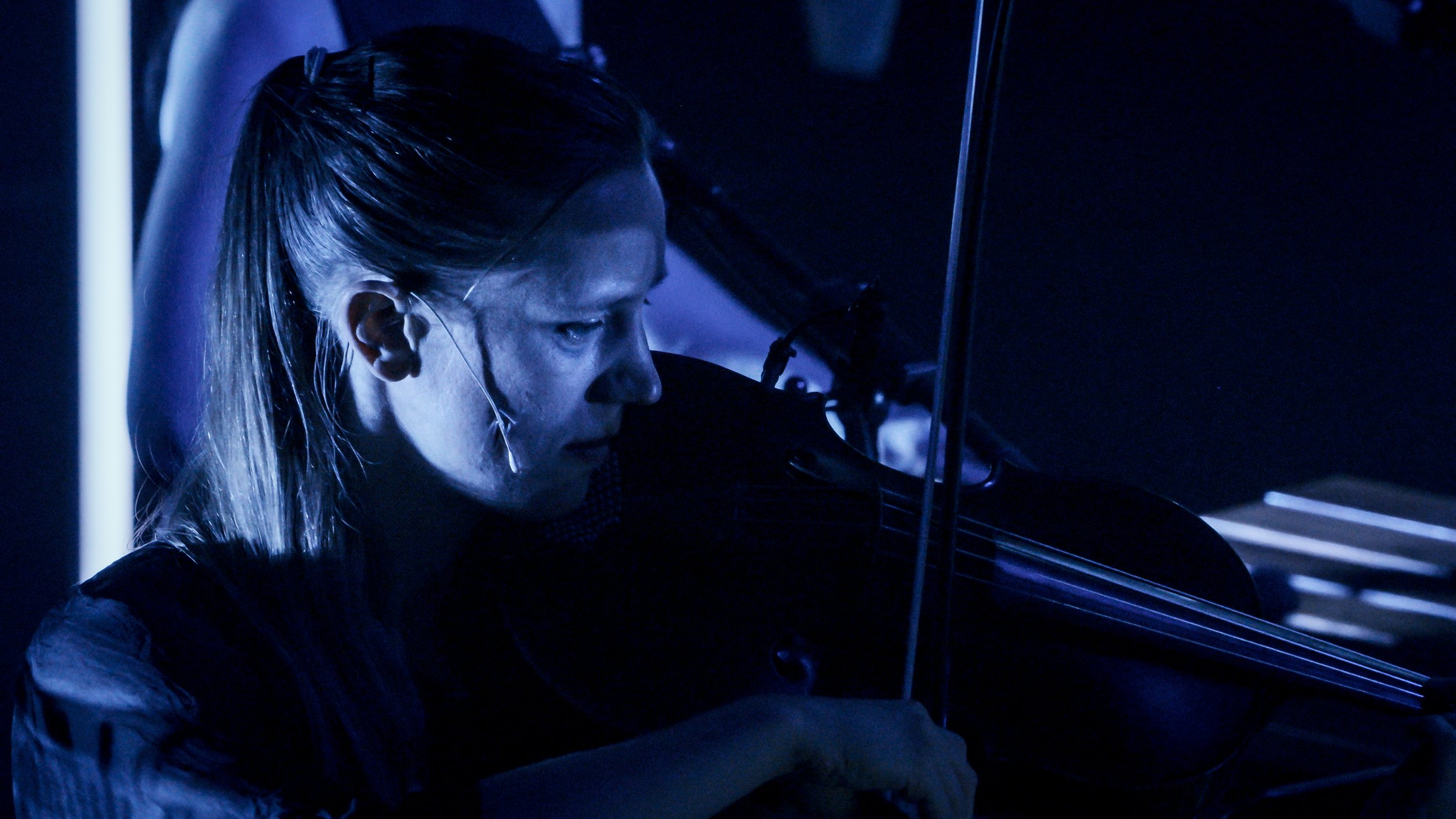 A woman with long, blonde hair playing a violin in a dimly lit environment with blue lighting.
