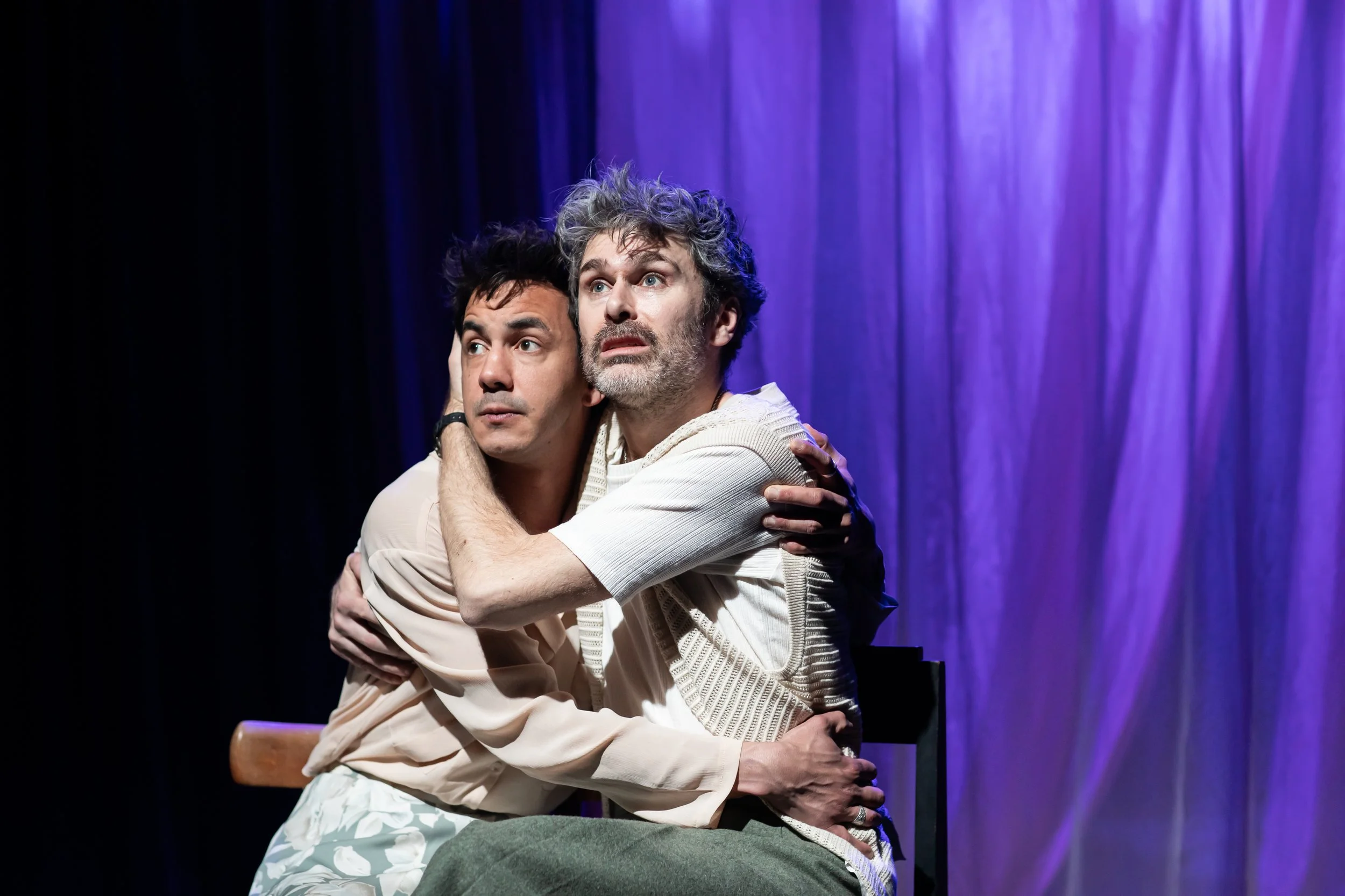 Two men hugging on stage with purple curtains in the background, appearing emotional