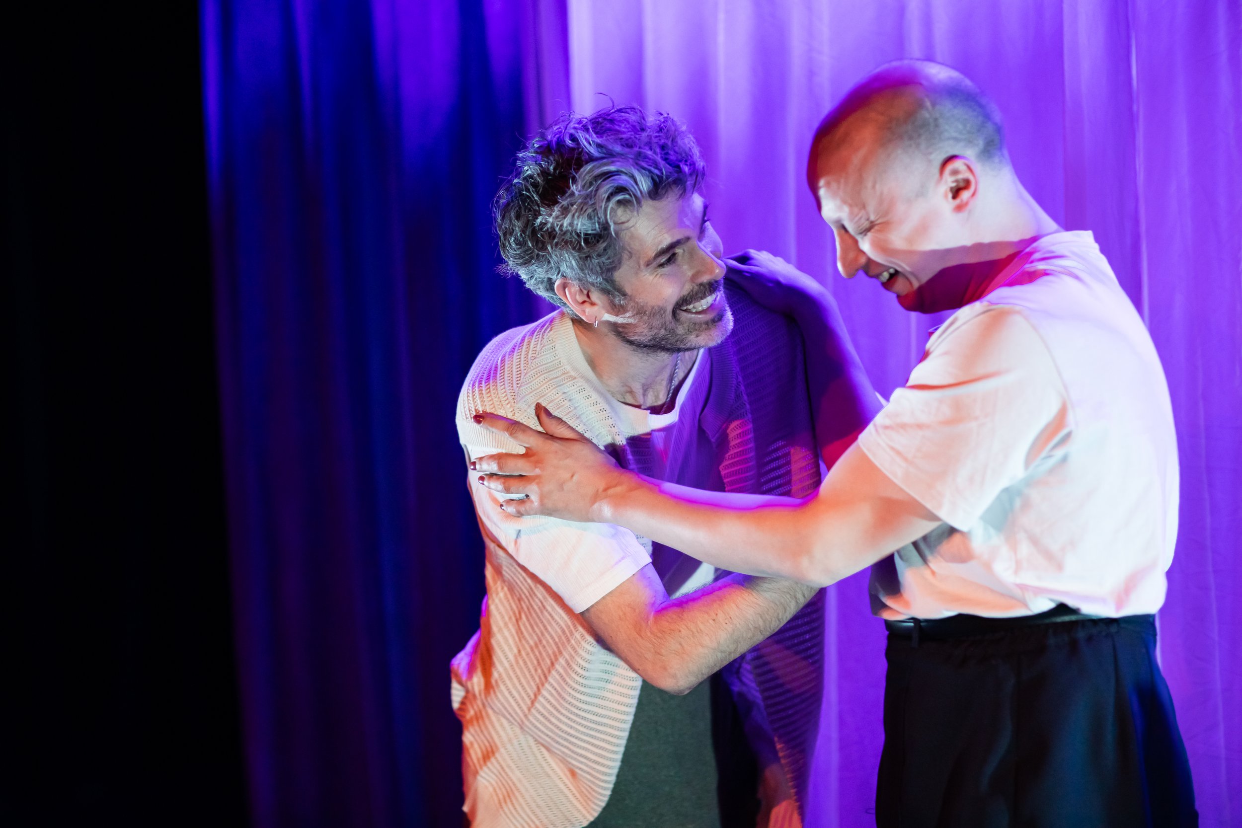 Two men are laughing and embracing each other on a stage with purple lighting, with a curtain in the background.