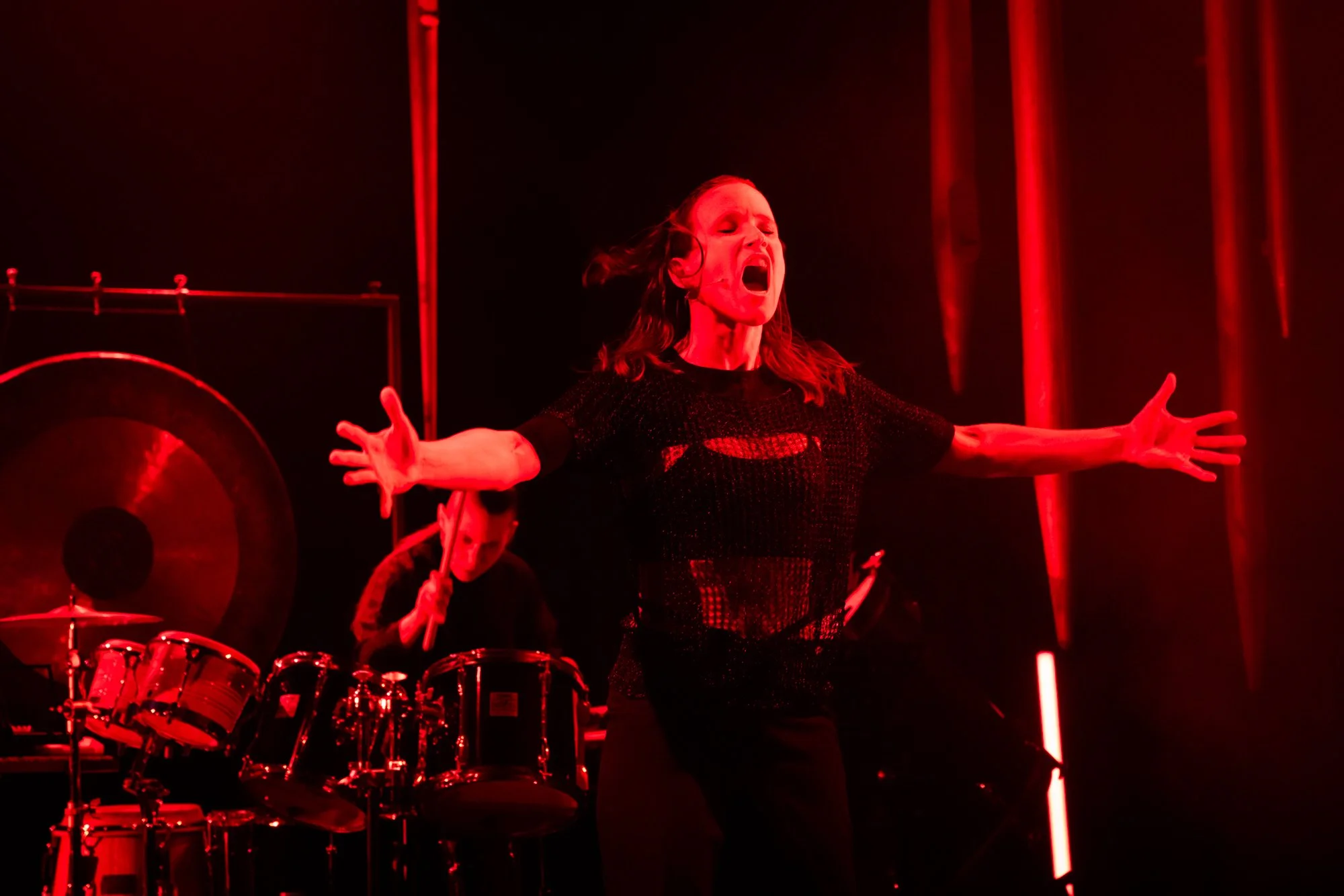 A woman passionately singing or performing on stage, with red lighting, in front of a drum set and gong, wearing a black distressed shirt.