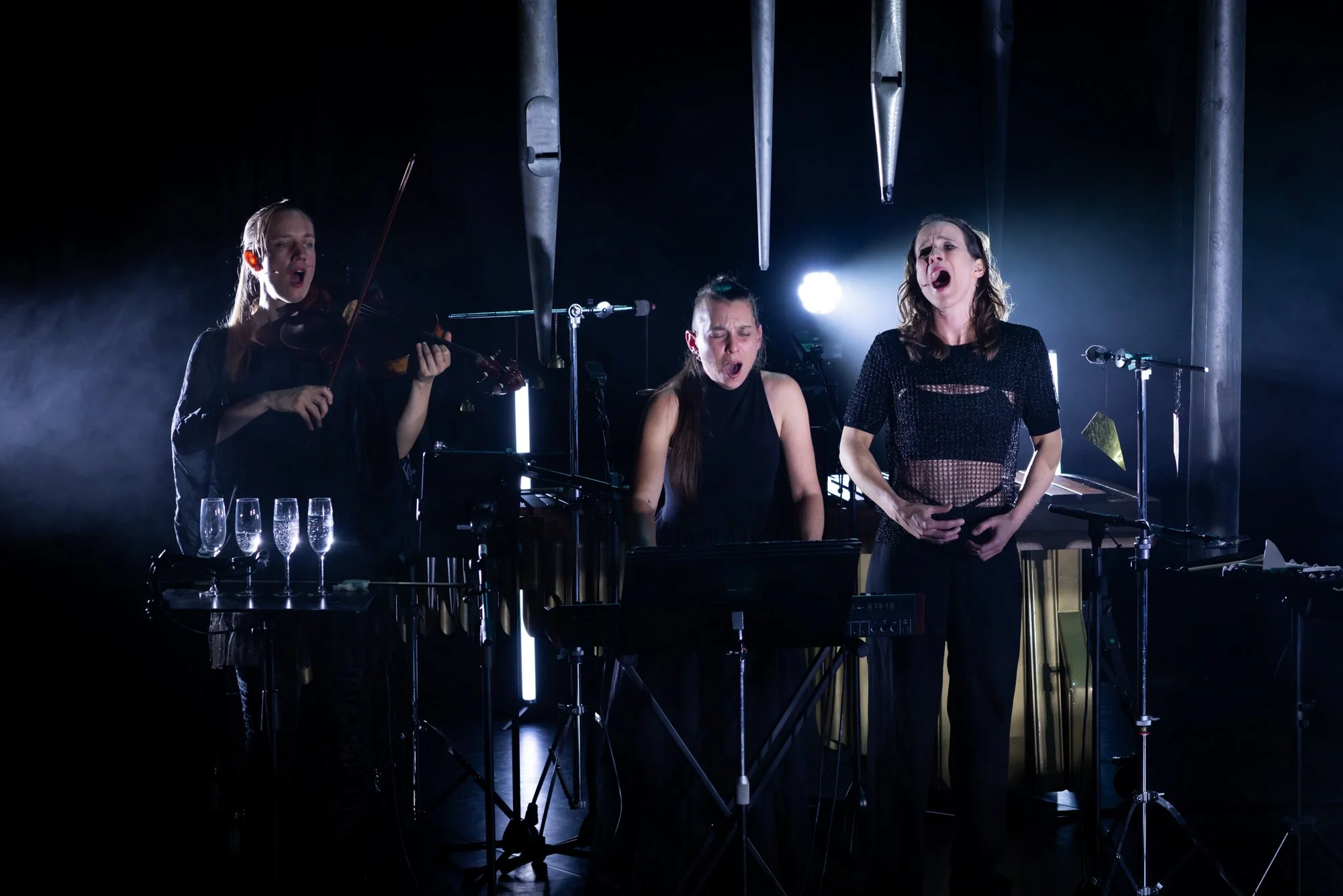 Three women performing on stage with musical instruments and microphones, one playing the violin, others singing, with dramatic lighting and music equipment around.
