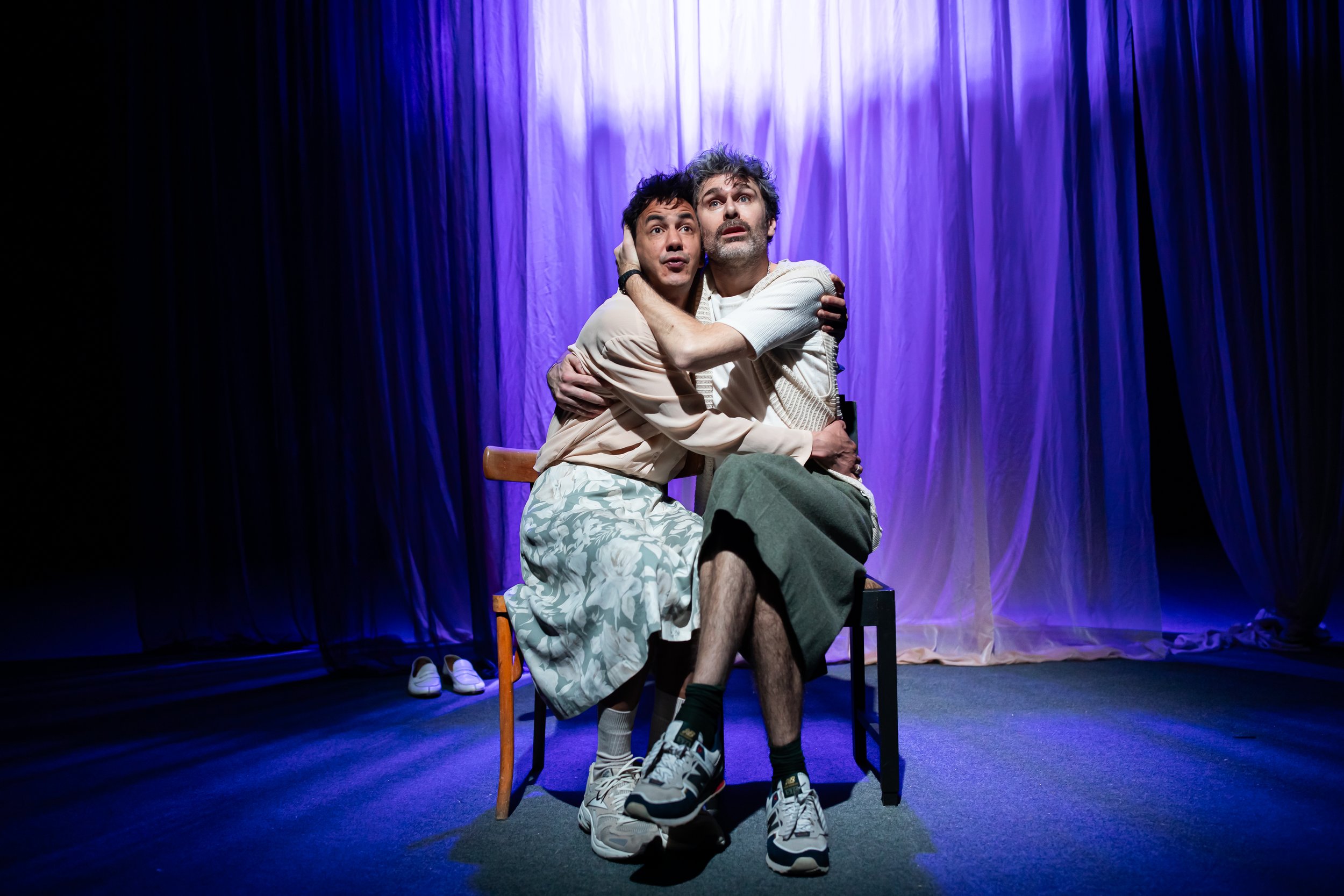 Two actors sit on chairs on stage, embracing and looking up, with purple curtains in the background.