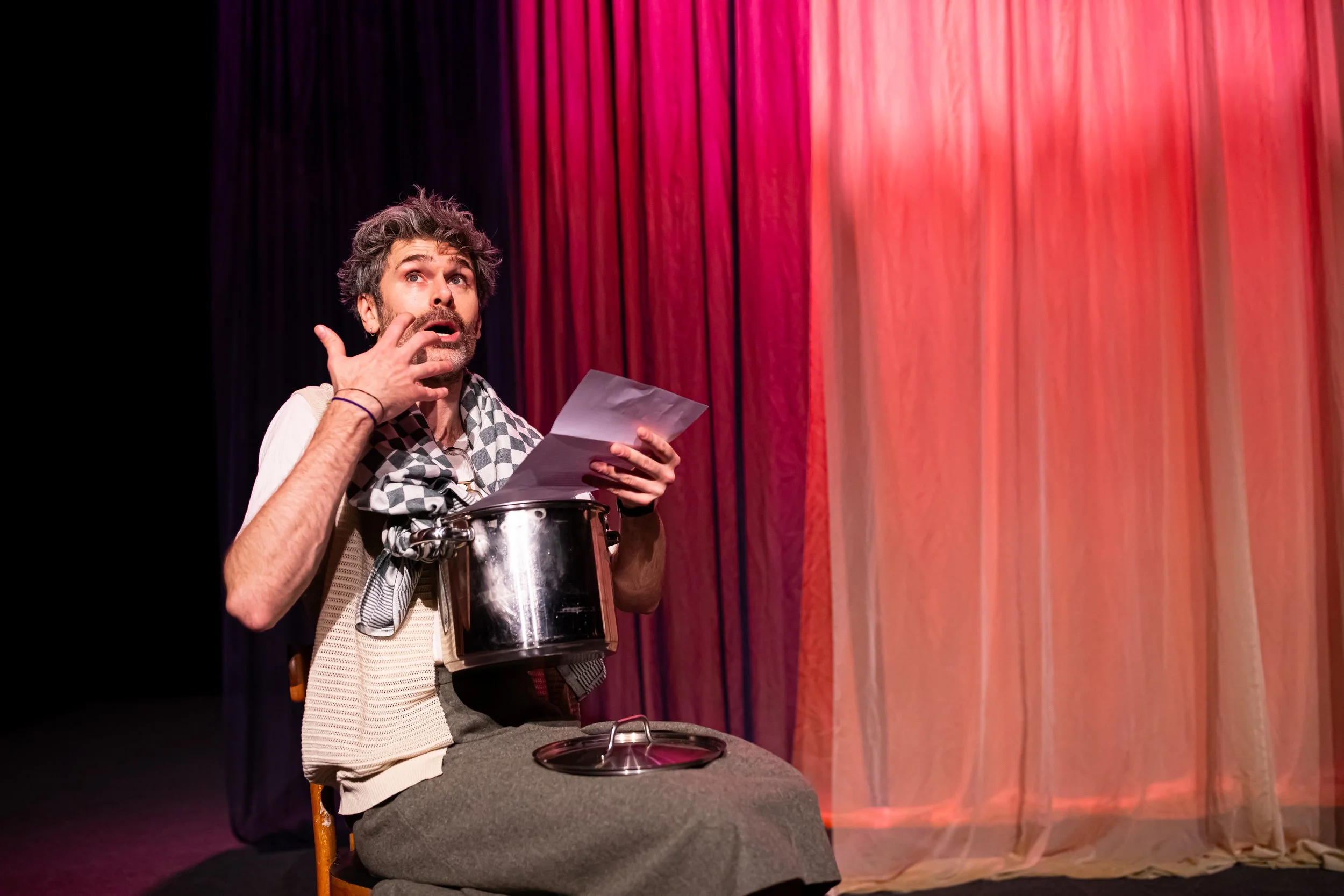 A man performing a theatrical monologue on stage, sitting on a chair with a pot on his lap, holding papers, with colorful curtains in the background.