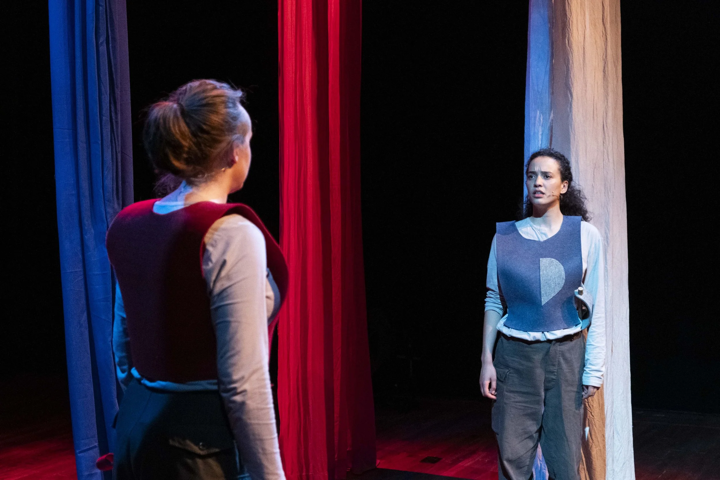 Two young women on stage, facing each other, with curtains in the background. One woman has her back to the camera, wearing a beige shirt and maroon vest, and the other is facing forward, wearing a blue vest over a beige shirt and gray pants. The sce